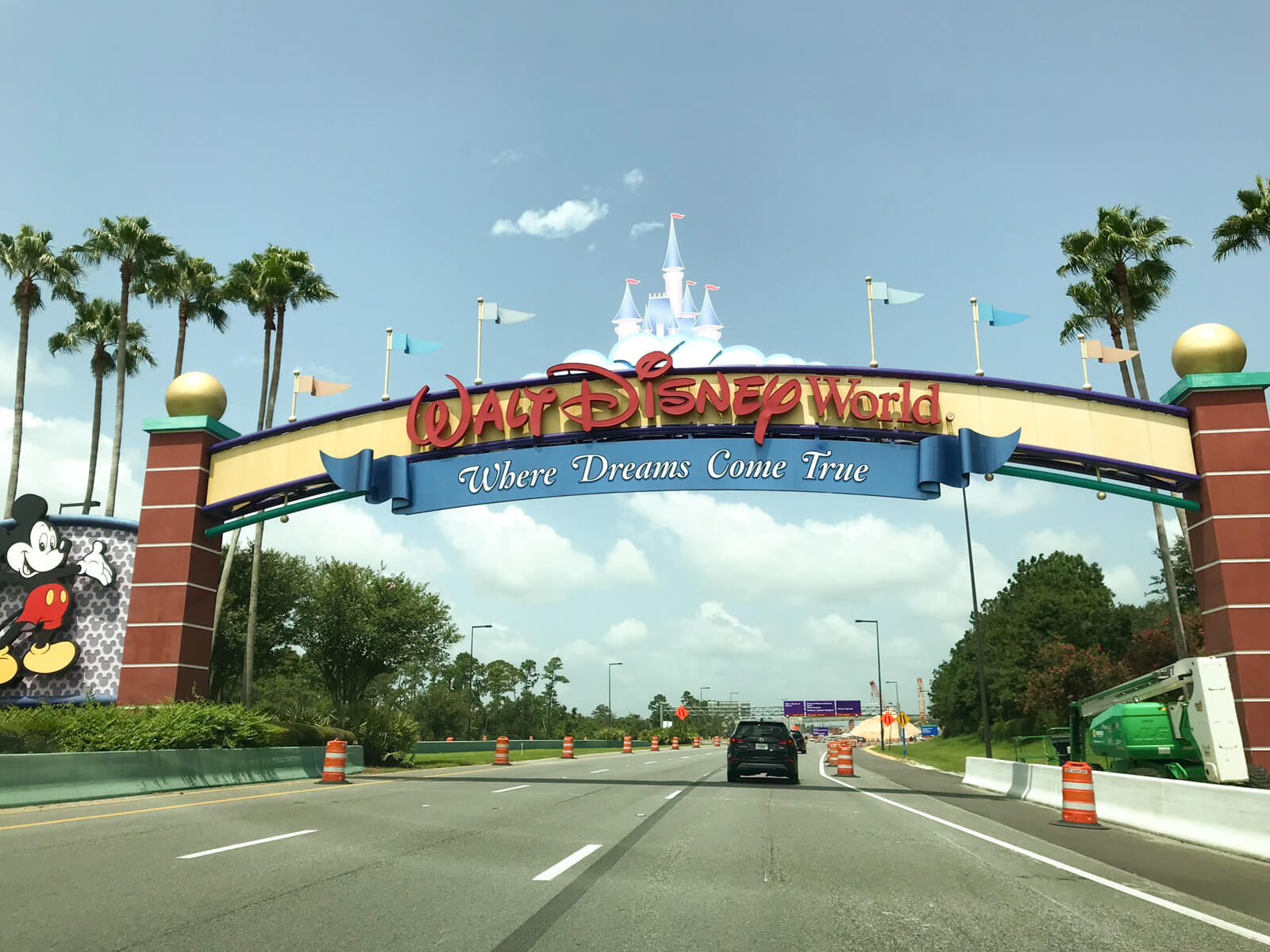 An arching sign with red text reading “Walt Disney World – Where Dreams Come True”. Several lanes of low traffic pass under the arch