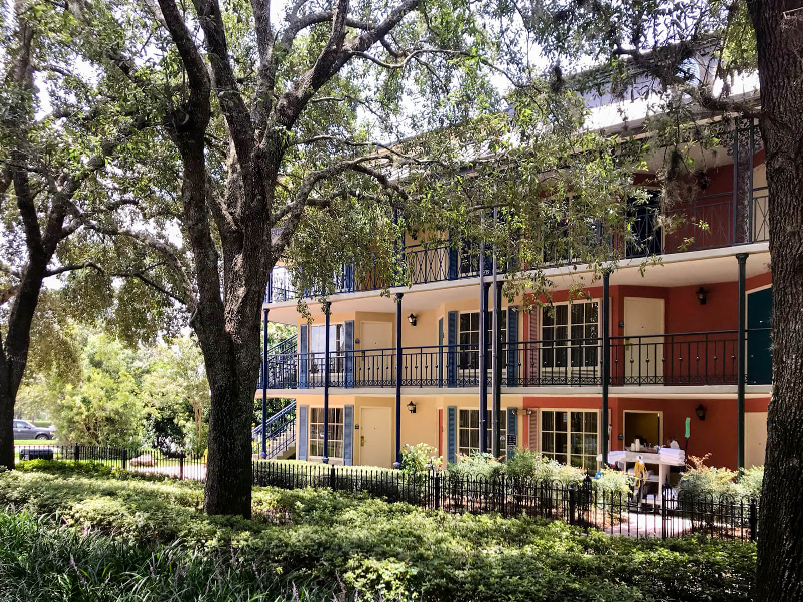 A building with three storeys of rooms. Each storey has a walkable area around the edge of the building. The building is painted brown-red, light yellow, and blue. In front of the building is a garden and some tall trees.