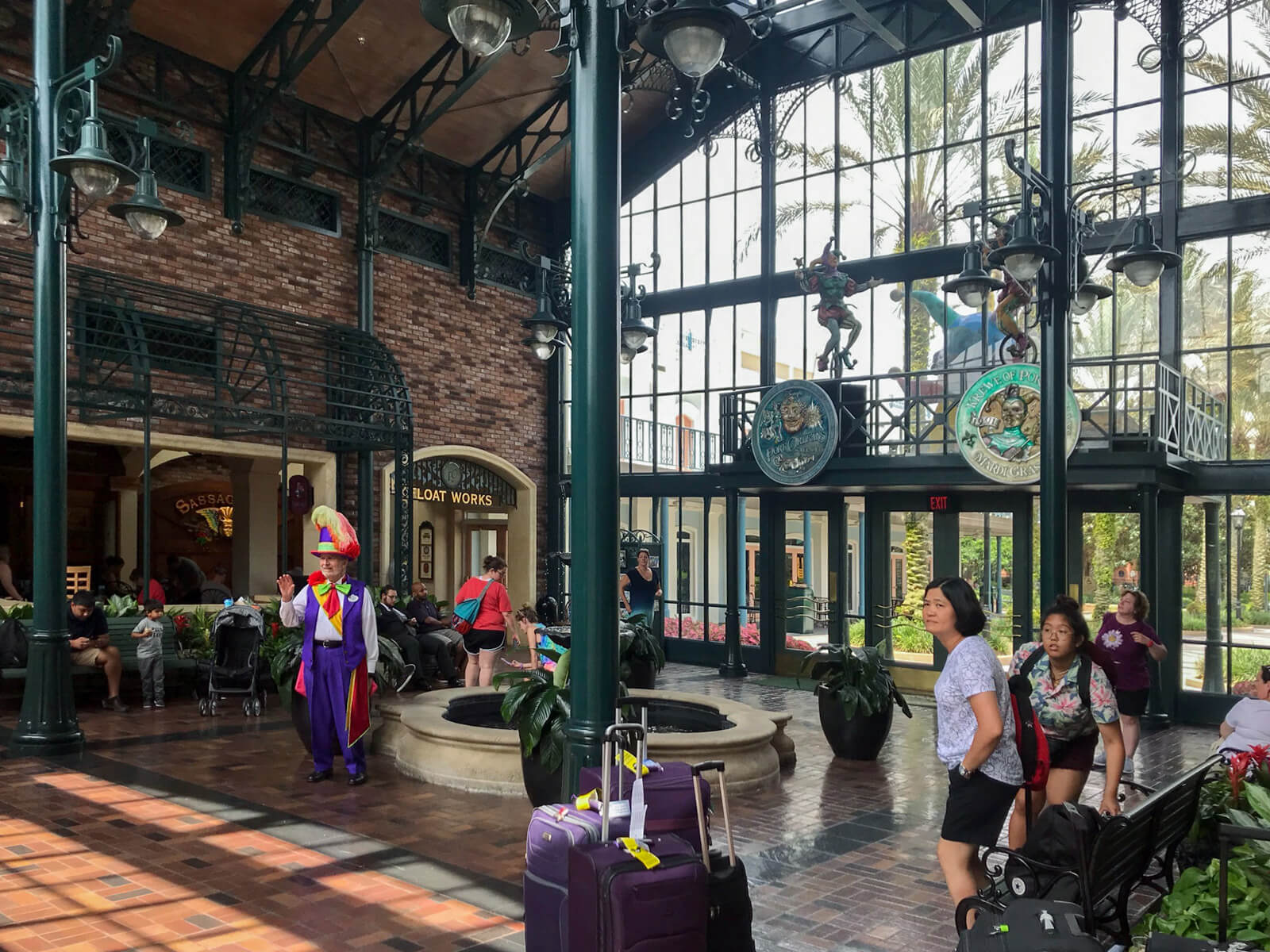 The inside of a lobby with green decor. There are people sitting on benches with luggage and prams. In the centre of frame is a small water fountain. To the right are glass doors letting a lot of natural light in.