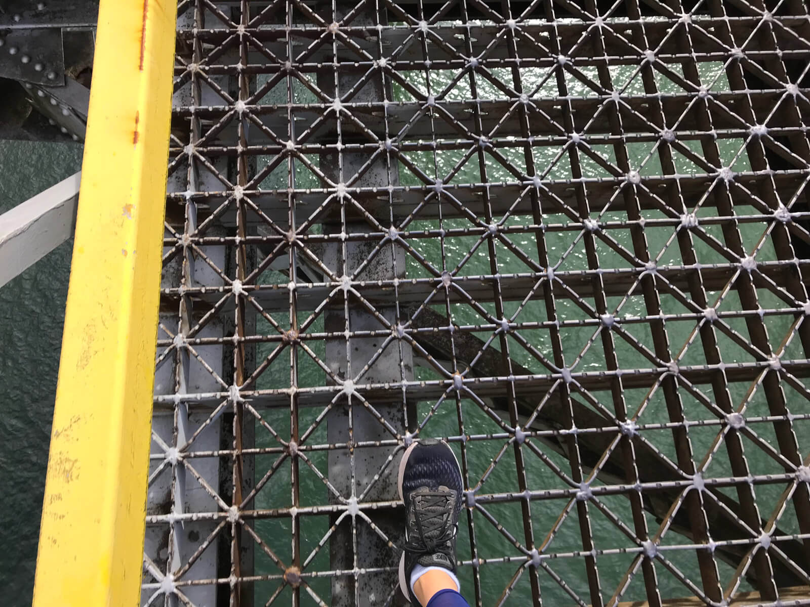 A birds-eye view of the walking surface of a bridge with water below. Someone’s foot, wearing running shoes, is in the frame. The holes between the bridge structure look quite large.