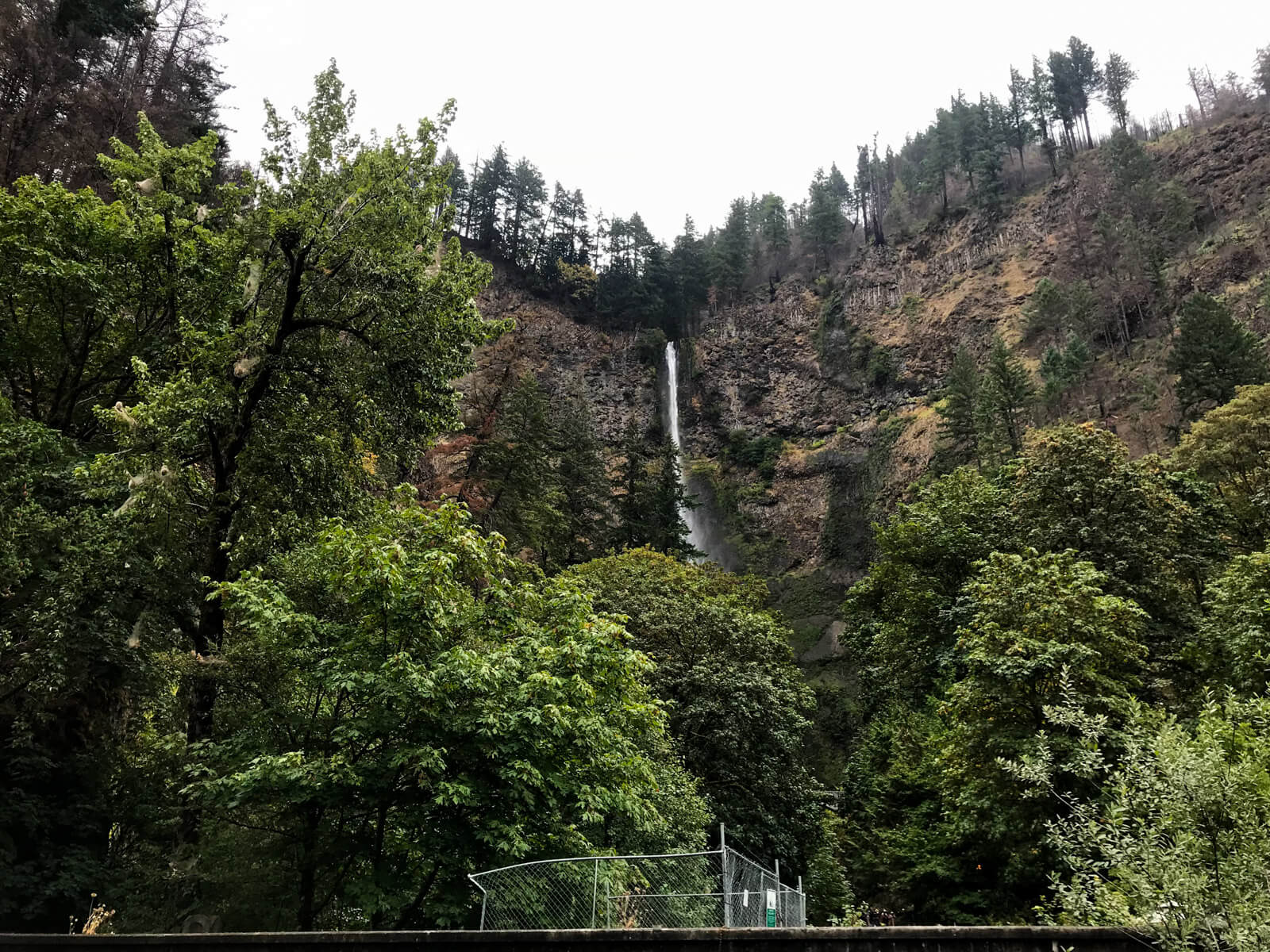 A low angle view of a tall waterfall. In the foreground is a bit of construction fencing