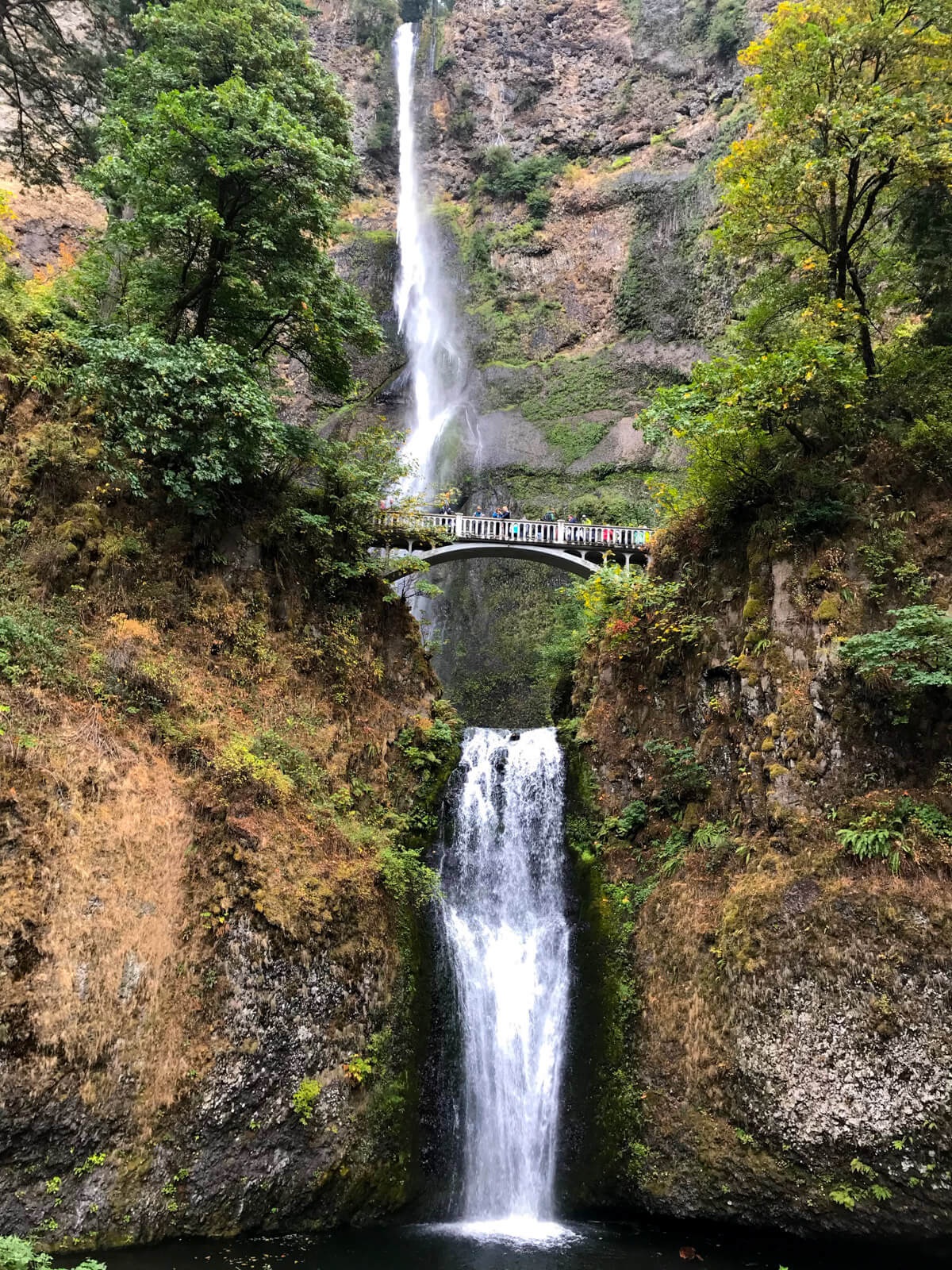 A narrow waterfall with brown rock and trees on the sides of the stream of water. The waterfall breaks in the middle where there is a small bridge, going over the width of the waterfall, with people standing on it