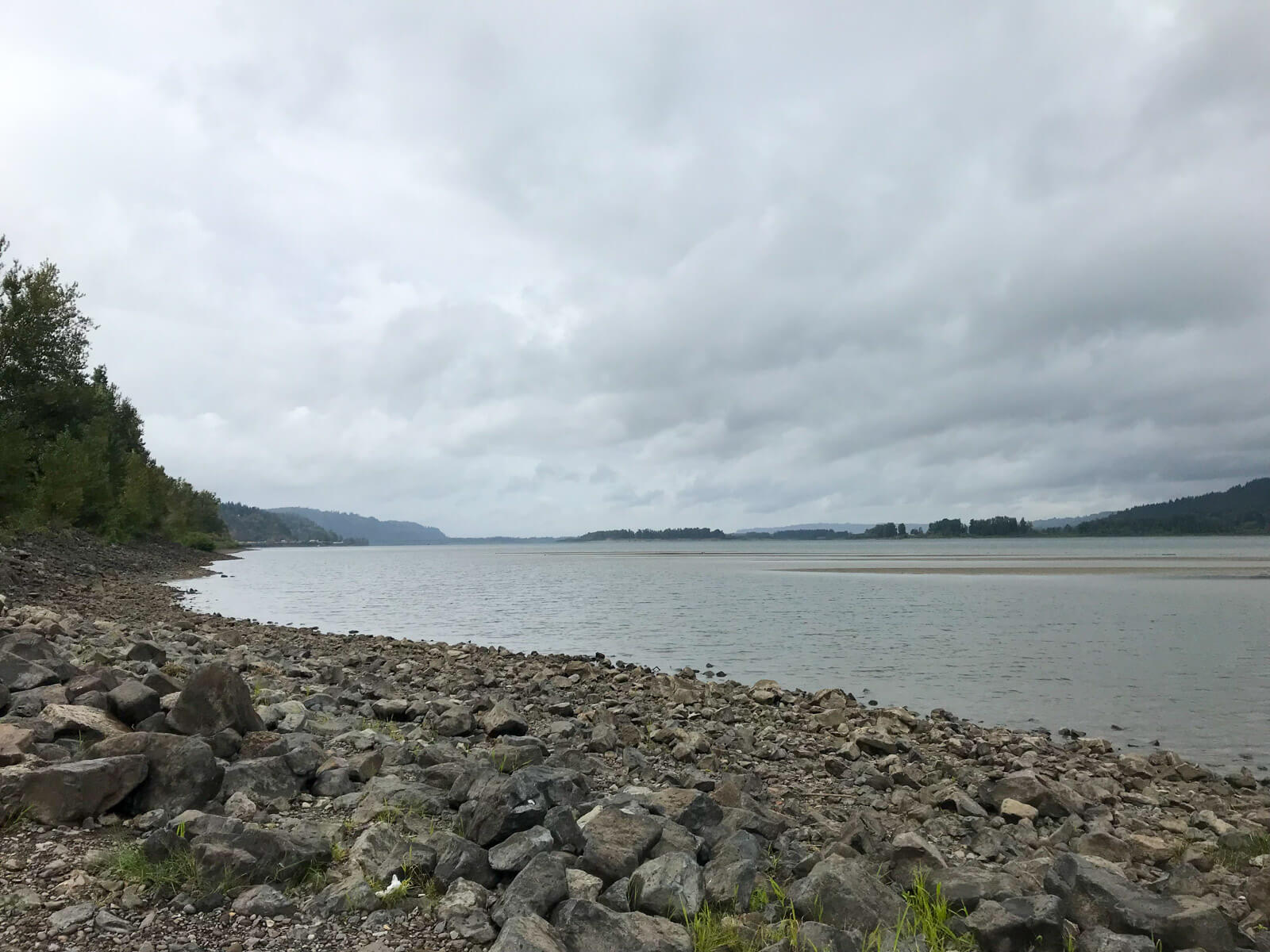 A rocky area leading to shallow water. It’s a cloudy day. The water looks shallow up until a patch of sand in the distance.