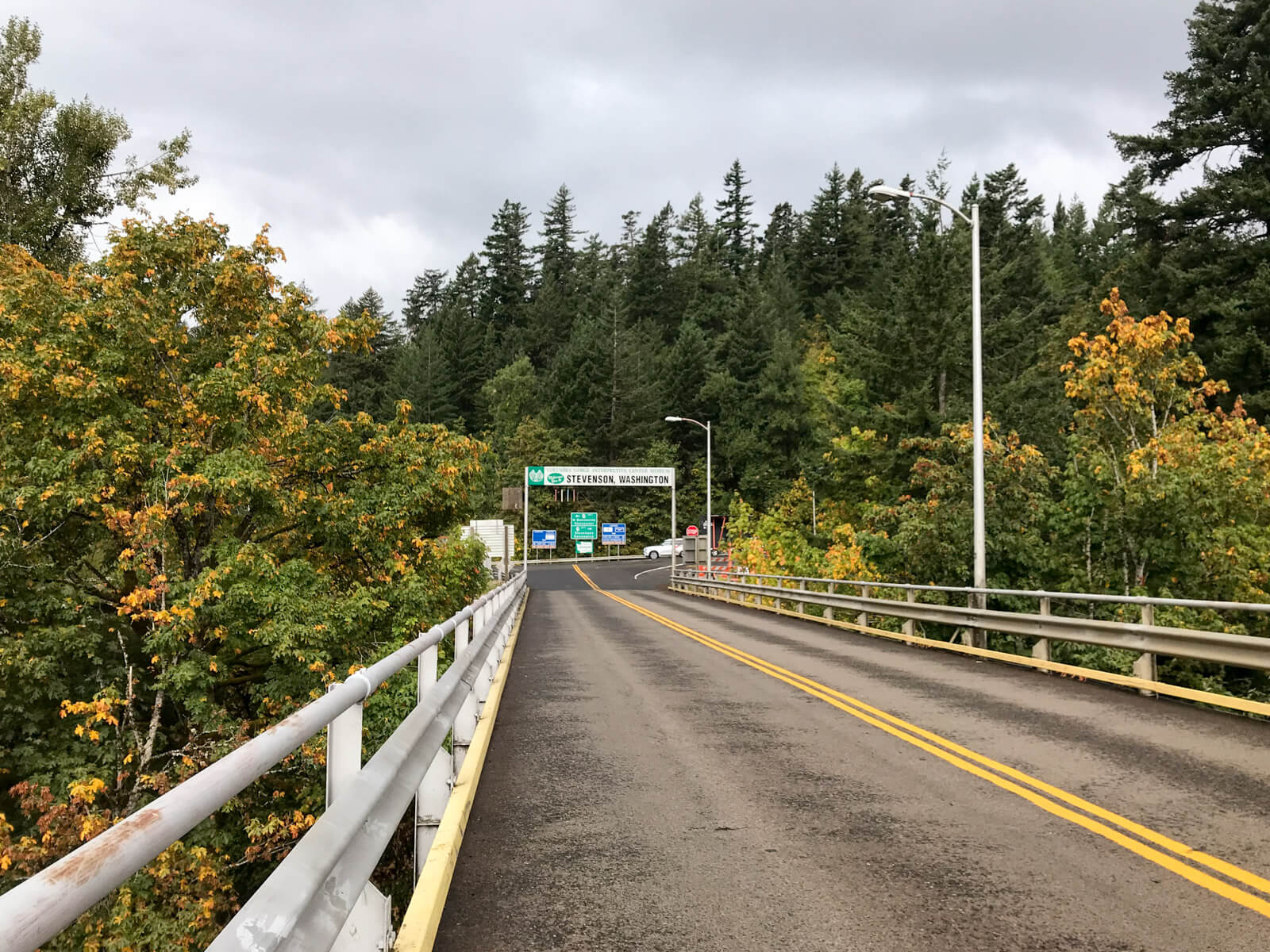 A view on an elevated highway bridge, with a sign ahead reading “Stevenson, Washington”, above the height of traffic. In the background are many green trees