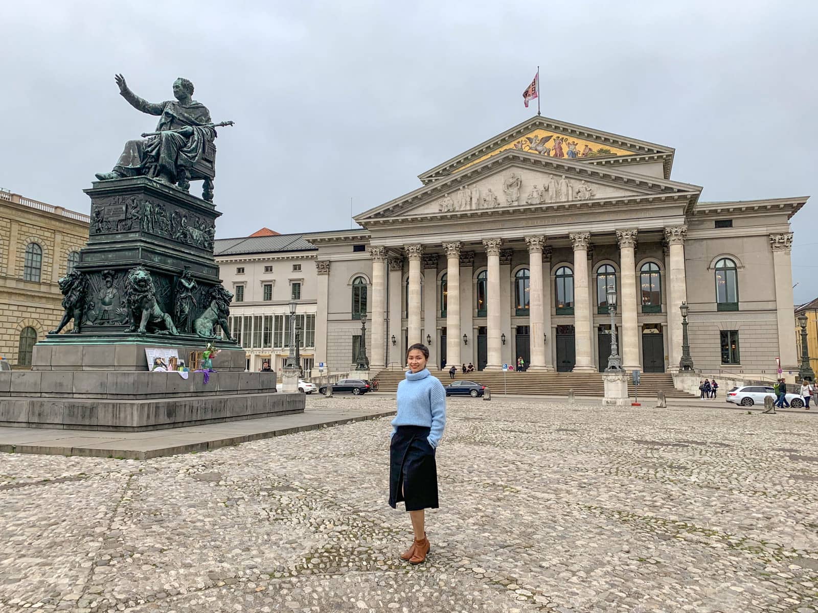 A woman in a blue sweater and black skirt in the foreground, she’s standing in front of an old building in Munich