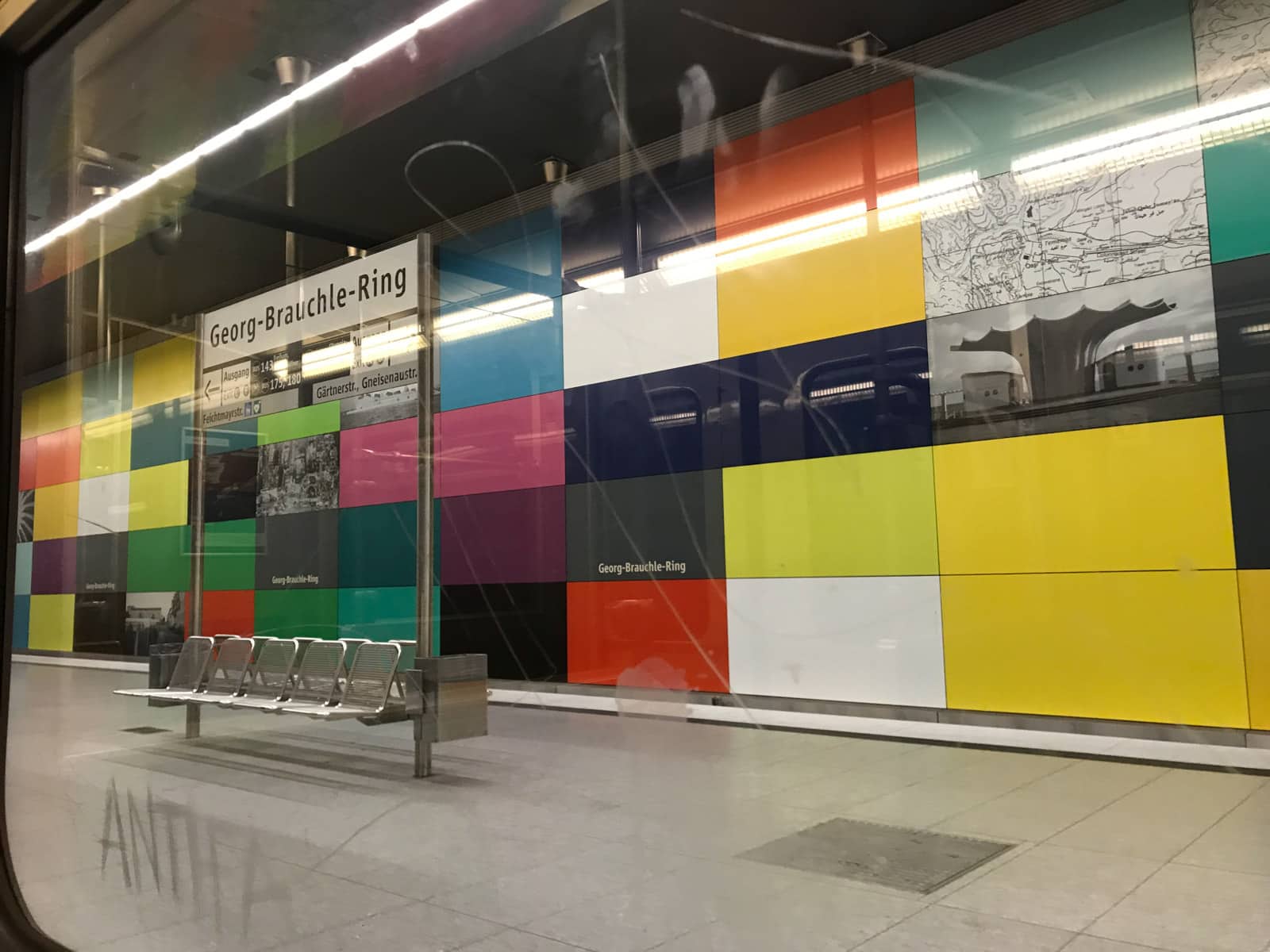 A view from the inside of a train window of a train platform. Five empty metal seats are on the platform, and the design on the walls consists of different coloured rectangular tiles, some with black-and-white photography