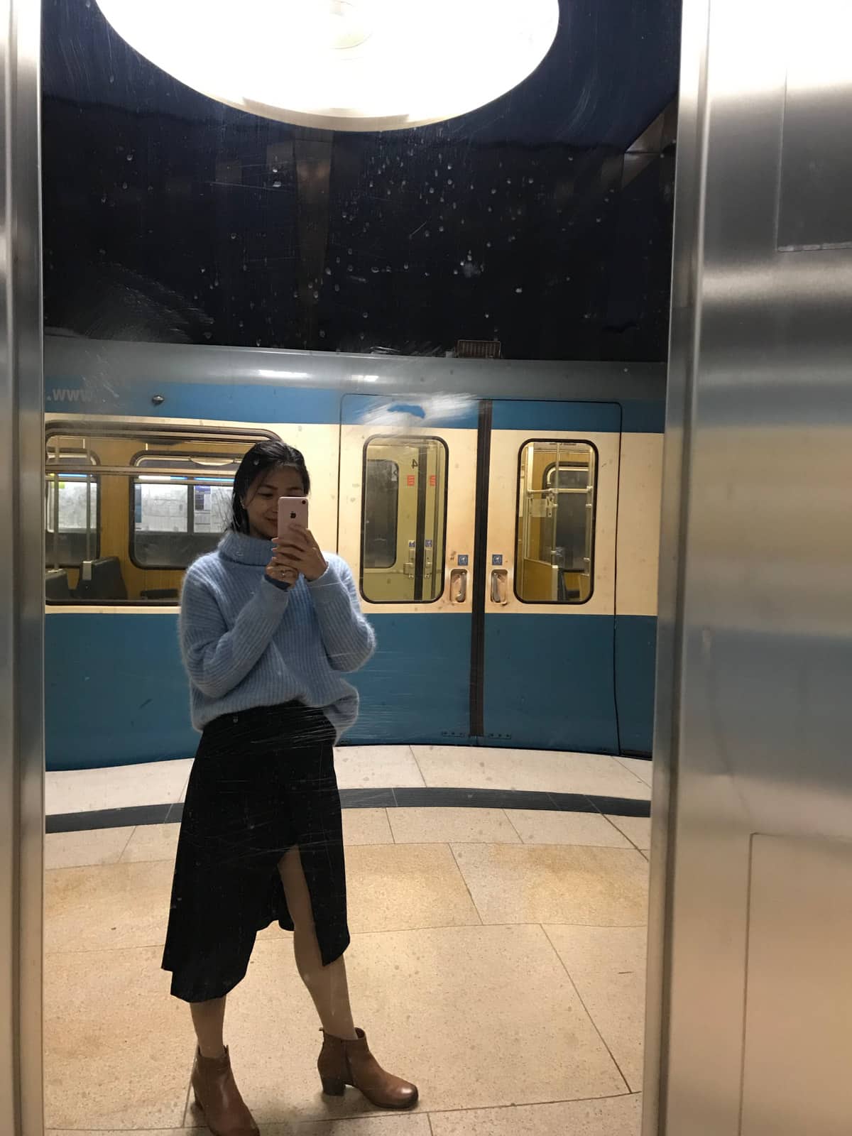 A woman in a blue sweater and a long black skirt, taking a selfie in a mirror at a train station. In the background is a blue train