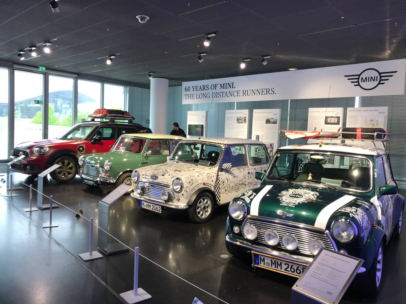 Four Mini Cooper cars in a museum, in a row, in different colours. A banner behind them reads “60 years of Mini. The long distance runners”.