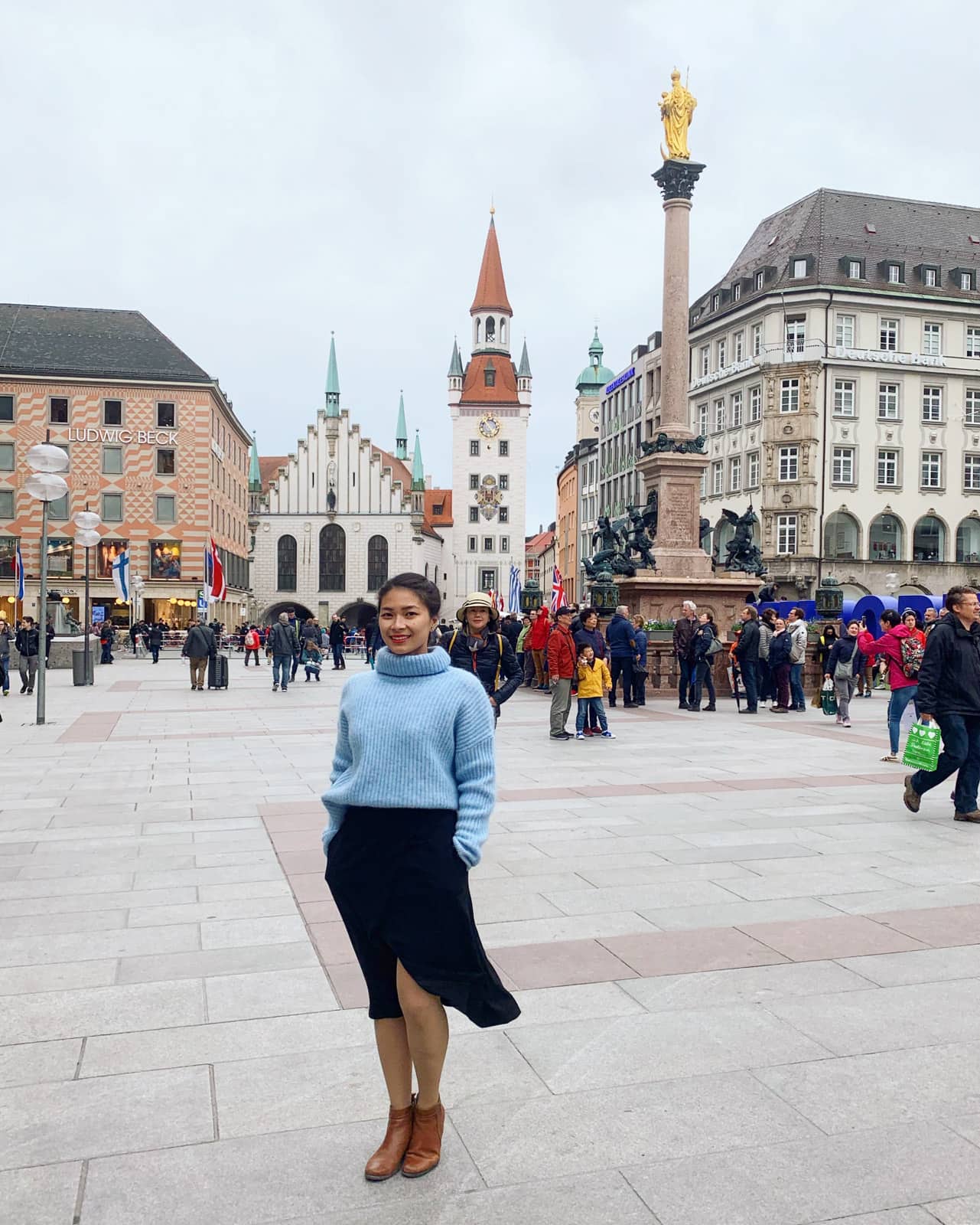 A woman in a blue sweater and black skirt in the foreground, she’s standing in a town square in Munich where there are many people in the background as well as old but pretty buildings