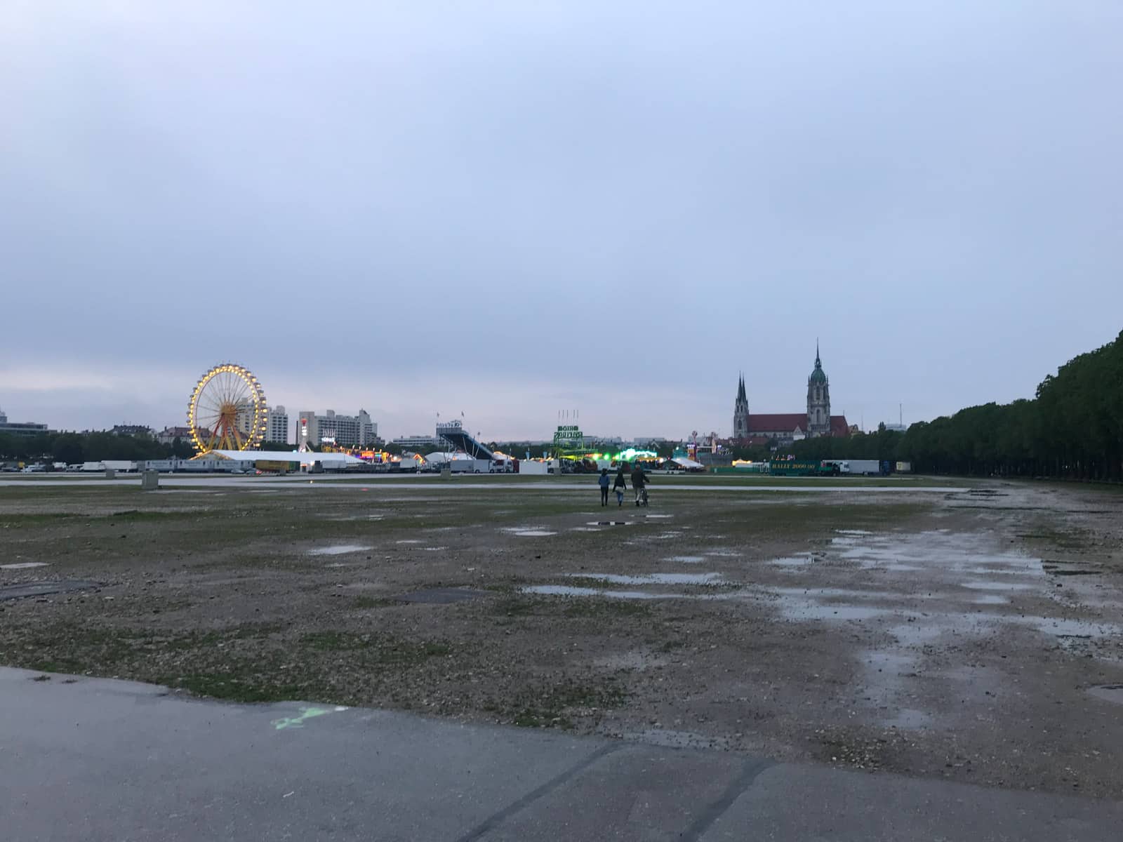 An open park area in the evening. The grass is wet from the rain. In the distance is a temporary festival ground set up with a ferris wheel and tents