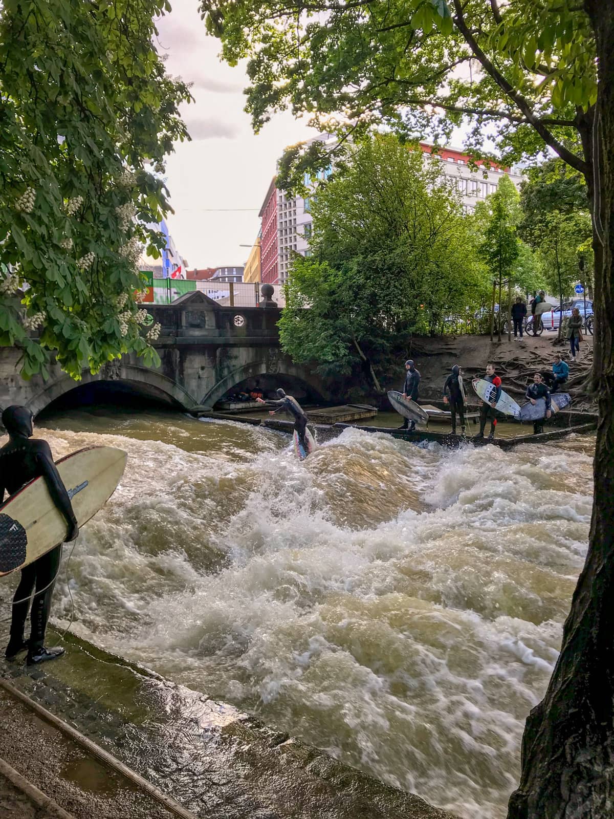 A section of waterway in a park, with high speed water running through it. Surfers in wetsuits are lining up to surf the waves, with one surfer already surfing