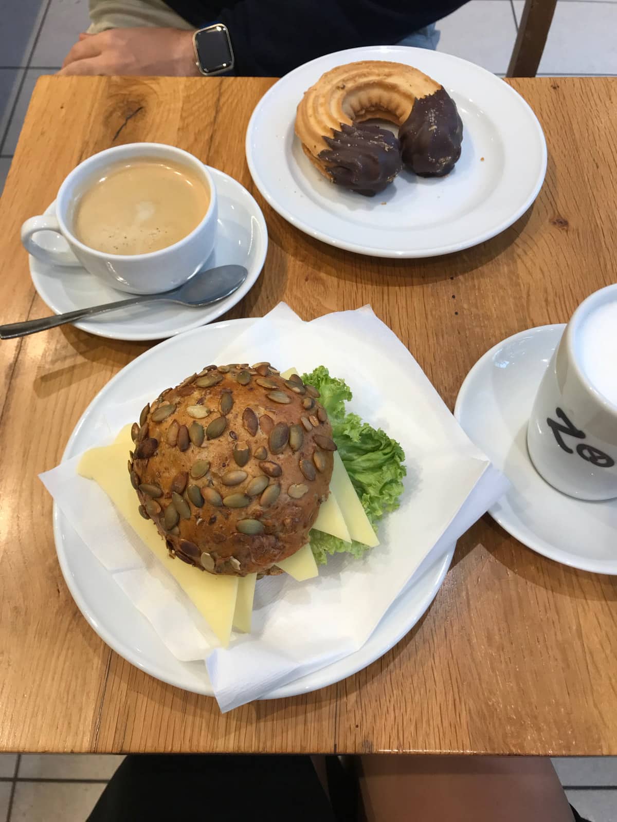 A small brown table with servings of coffee and two plates – one with a seed bun filled with cheese and lettuce, and the other with a chocolate dipped cookie