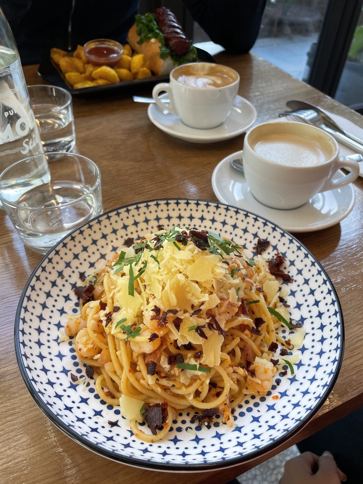 A bowl of spaghetti with chilli, prawn, and parmesan, on a table with some cups of coffee in the background