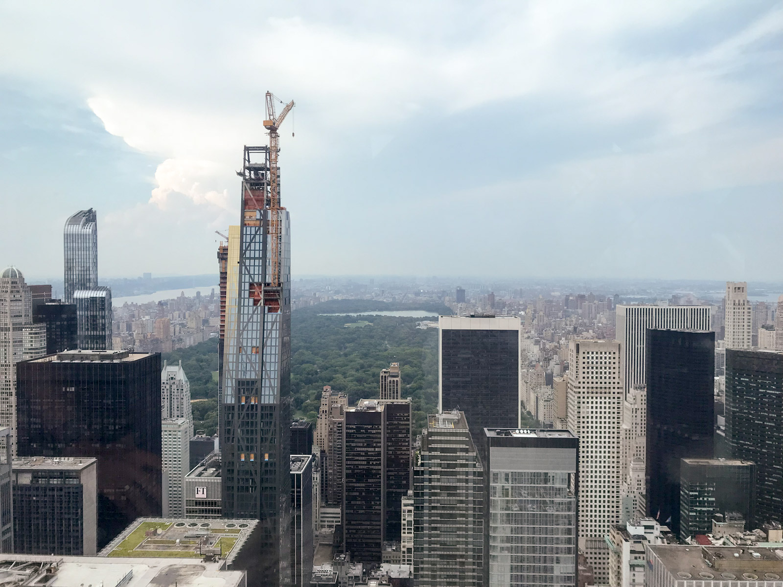 A view of a city with high-rise buildings. Some have active construction work and scaffolding. In the middle of the city is a large area showing a canopy of trees