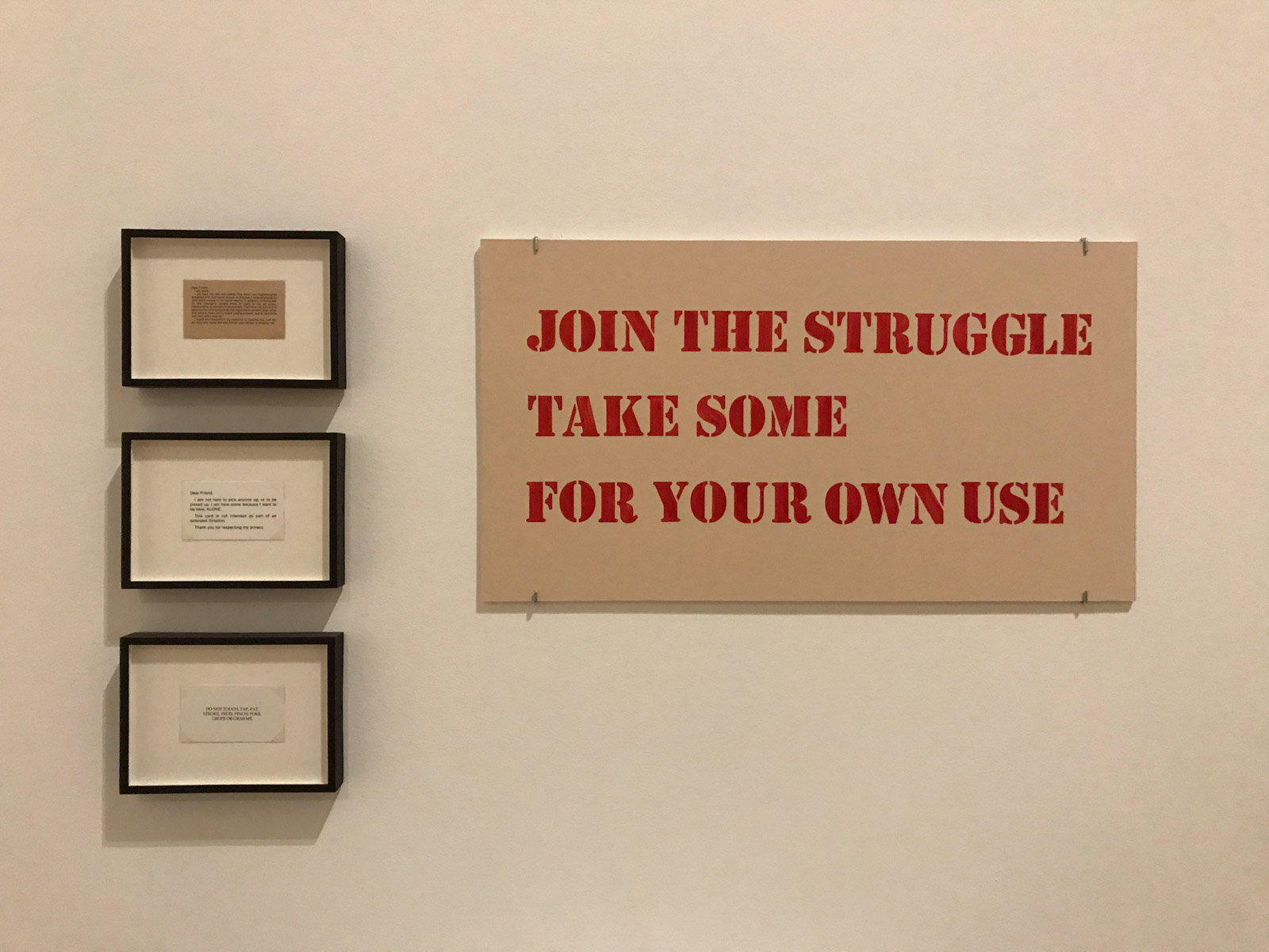 Three small cards in a vertical line, framed in glass with black border. To the right is a beige sign in red uppercase text, reading “Join the struggle. Take some for your own use”.