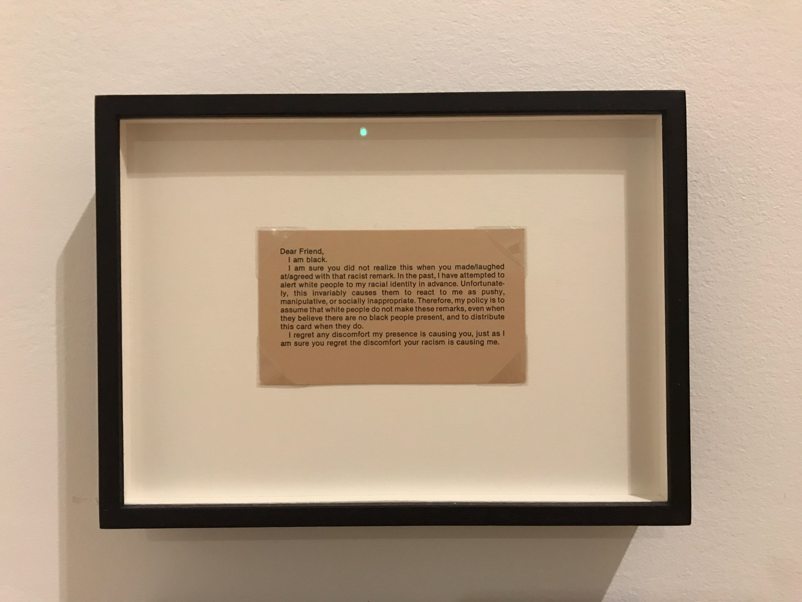 A framed brown card beginning with the following text: “Dear Friend, I am black. I am sure you did not realize this when you made/laughed at/agreed with that racial remark. In the past, I have attempted to alert white people to my racial identity in advance. Unfortunately, this invariably causes them to react to me as pushy, manipulative, or socially inappropriate. Therefore, my policy is to assume that white people do not make these remarks, even when they believe that there are no black people present, and to distribute this card when they do. I regret any discomfort my presence is causing you, just as I am sure you regret the discomfort your racism is causing me.”