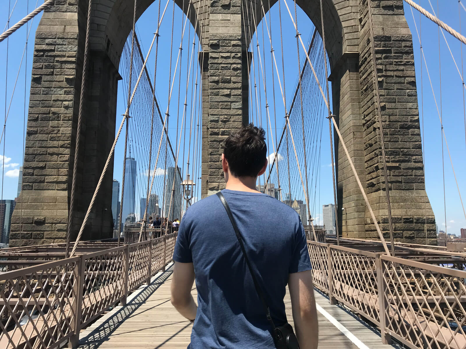 A man, from behind, wearing a blue shirt. He is walking a bridge with giant pillars of structure ahead of him