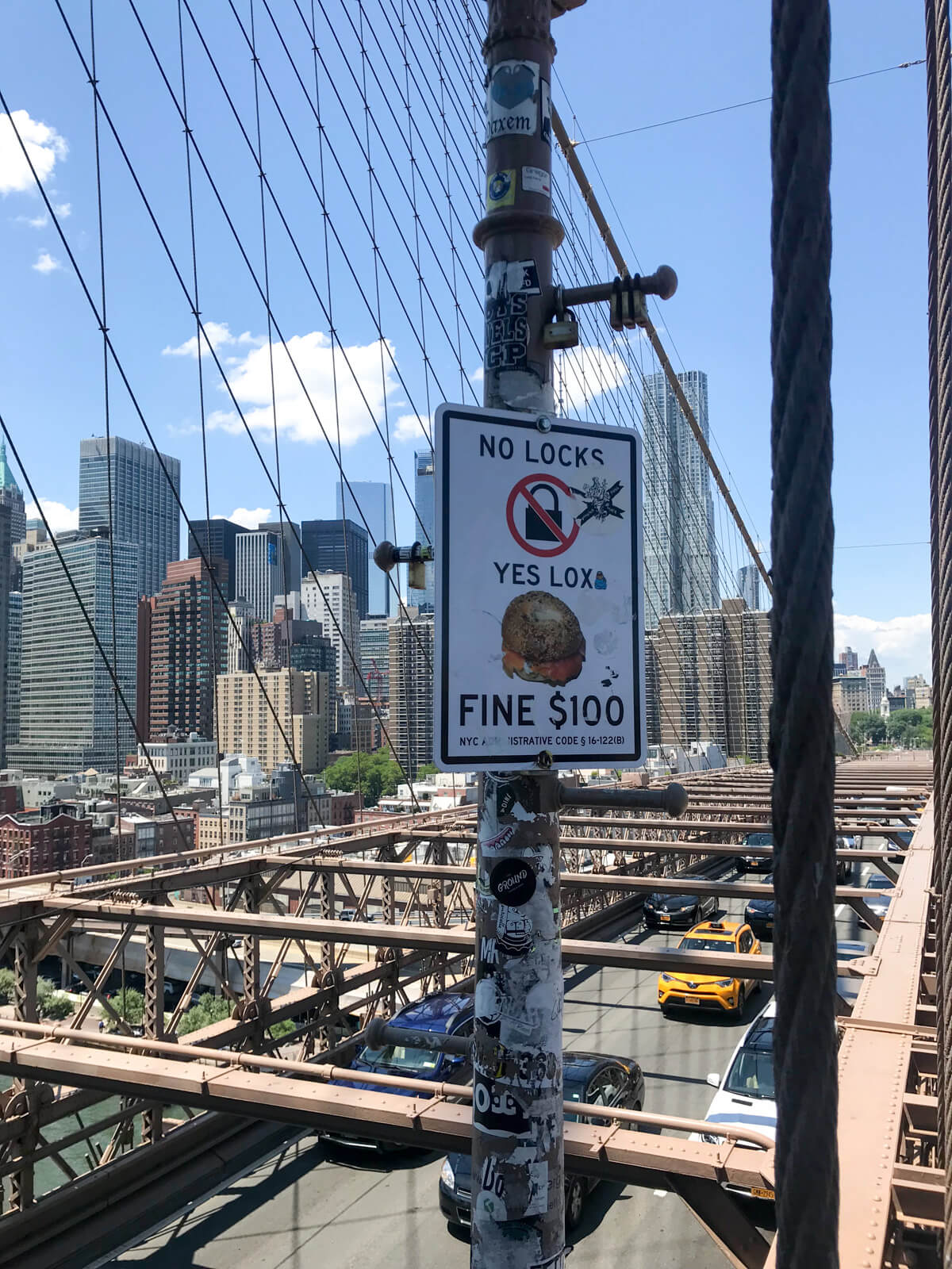 A sign on the structure of a bridge reading “NO LOCKS, YES LOX, FINE $100” showing an icon of a lock crossed out, and a burger with smoked salmon