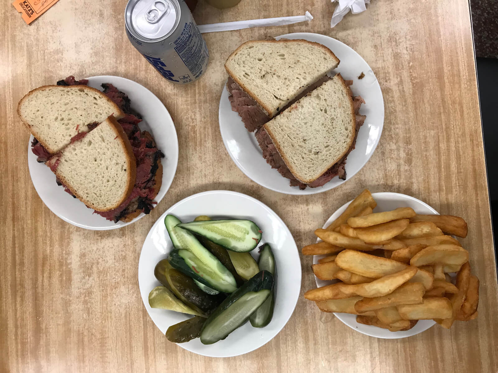 Several white plates on a wooden table – two with sandwiches cut in half, one with green pickles and the other with fries. There is also a can of Pepsi on the table