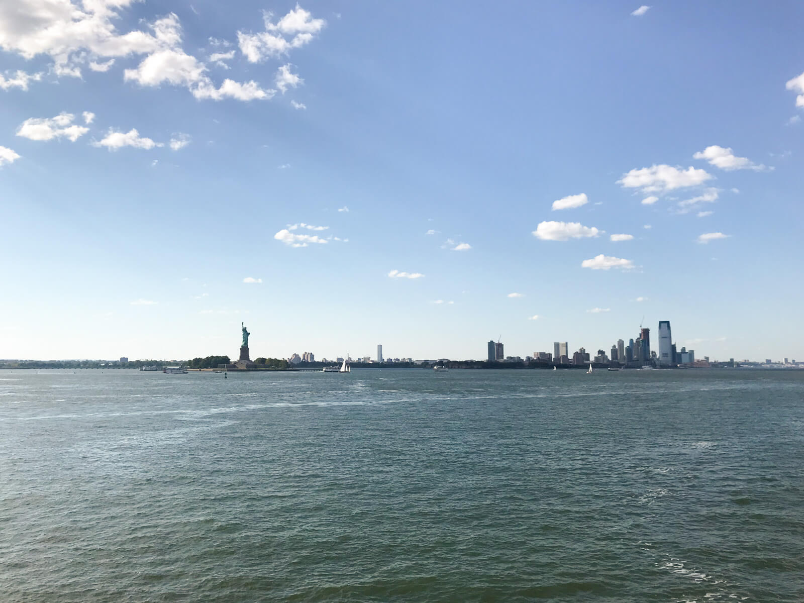 New York City with high-rise buildings and the Statue of Liberty as seen from the water on a ferry. The sky is a dim blue with some clouds to the left of frame.