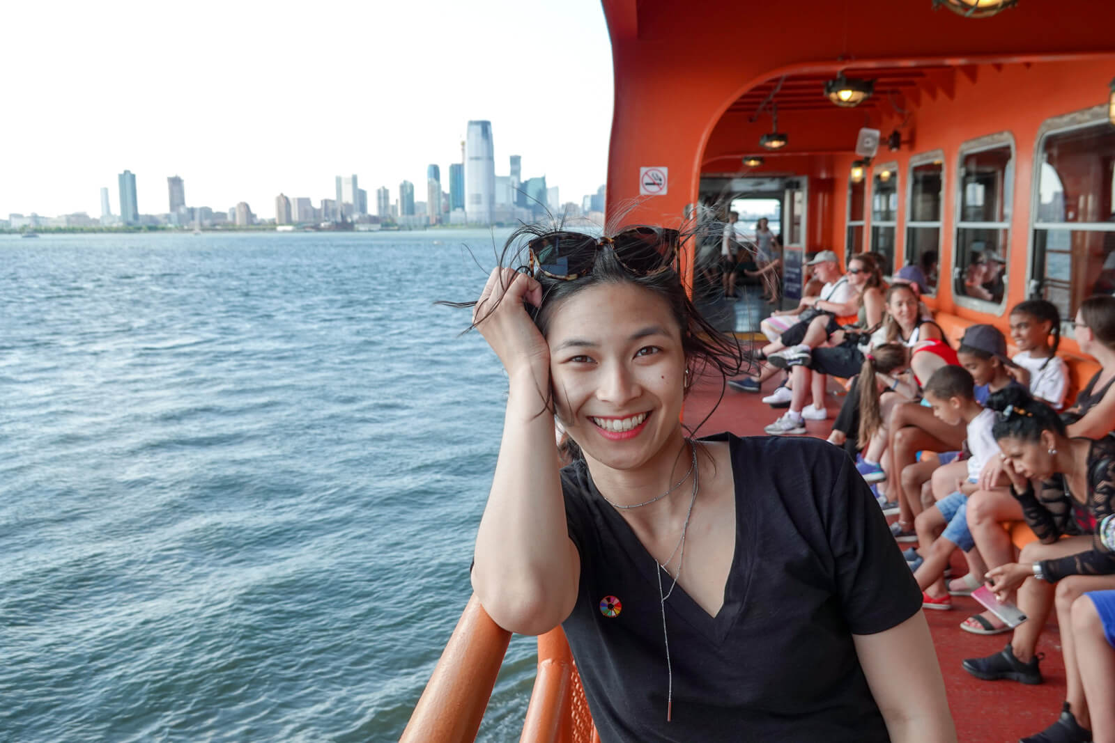 A photo of a woman on a ferry, her elbow leaning on a handrail. In the background is a New York City skyline