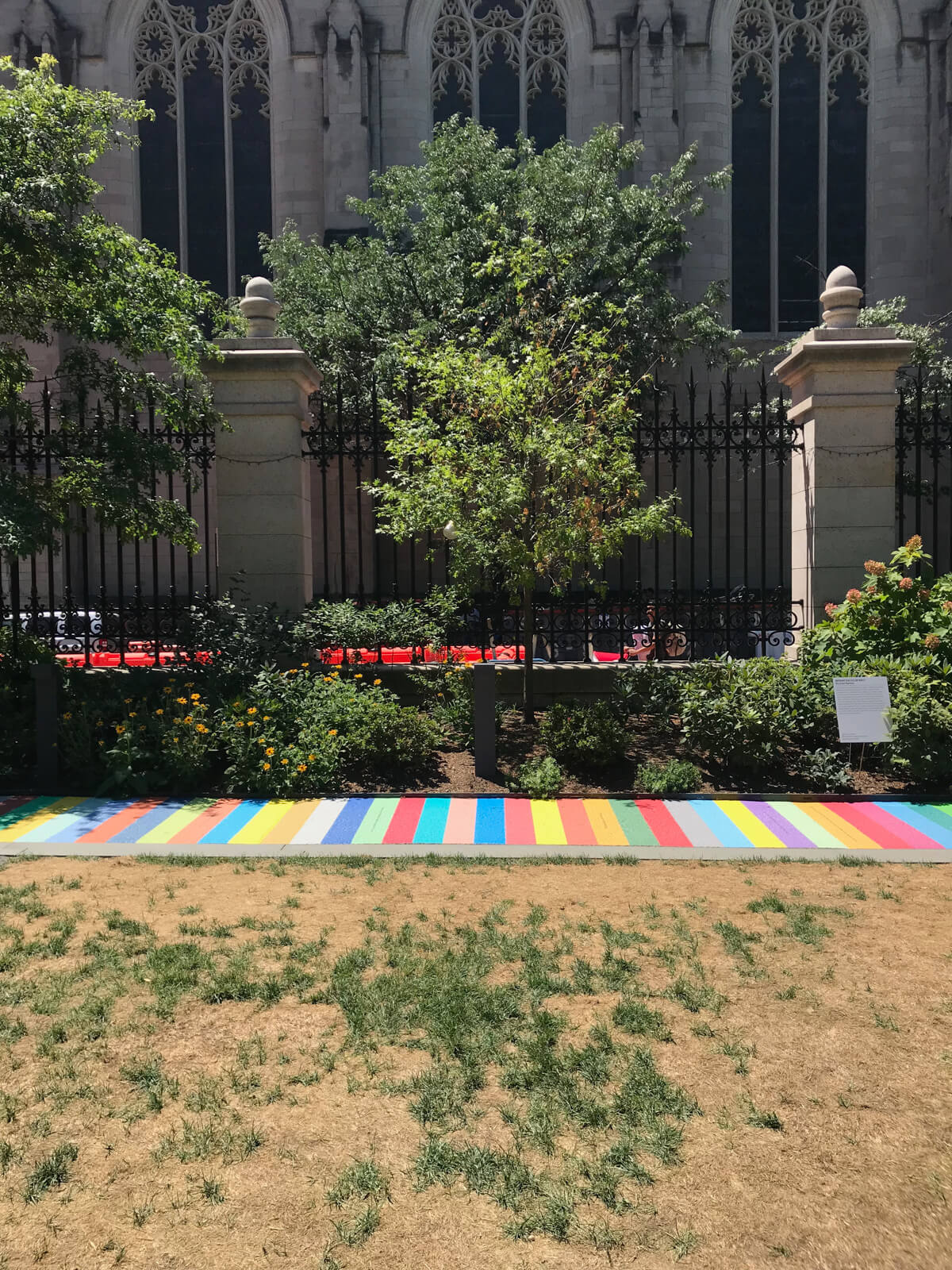 The inside of a park, walled off by pillars and black fencing. Towards the fence, from left to right, is a painted path (a walk) with different coloured stripes
