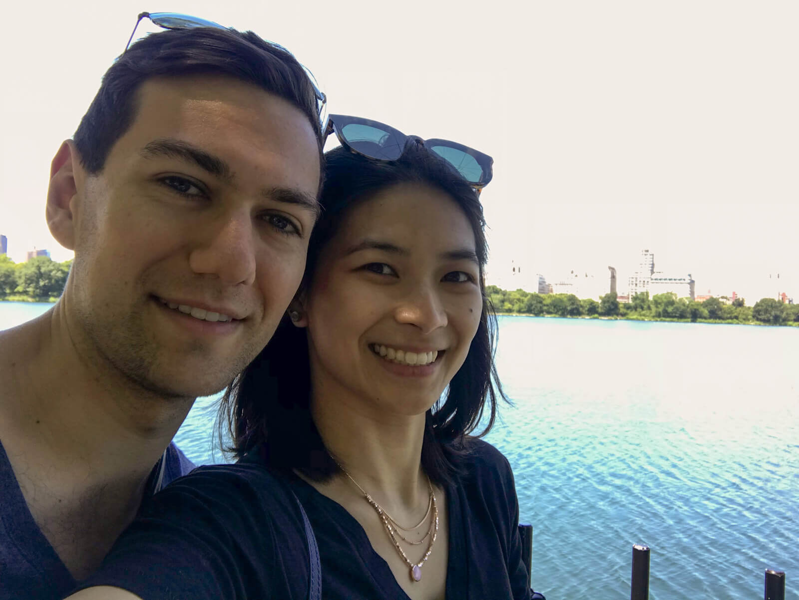 A man and woman smiling, both wearing sunglasses on their head. In the background is an overexposed lake