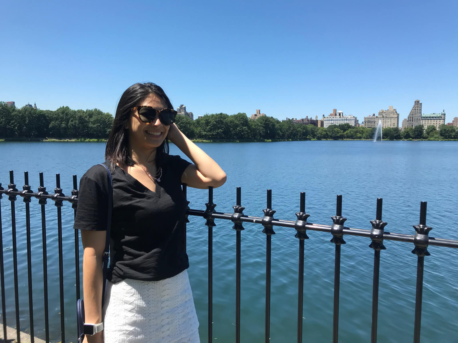 A woman wearing sunglasses, a black t-shirt and white skirt, standing by a black fence. In the background is a large lake and buildings and trees further into the distance