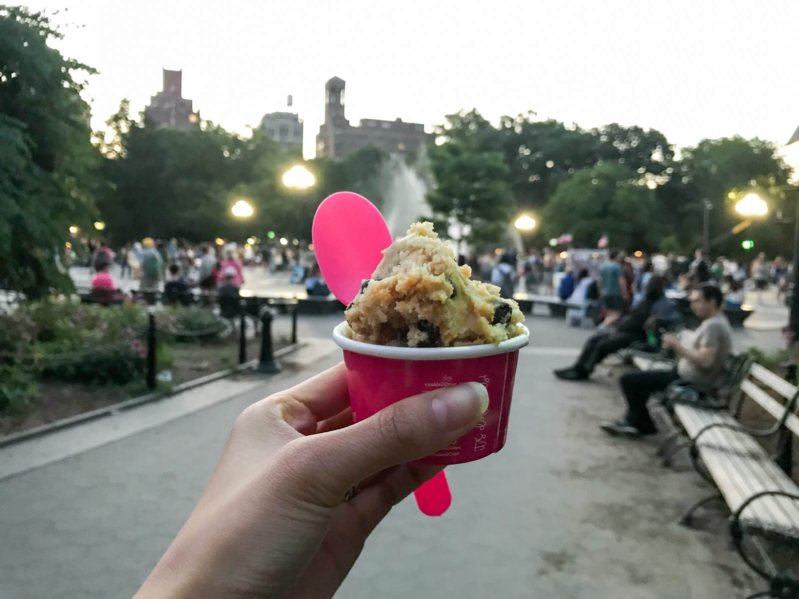 A person’s left hand holding a small pink cup and pink spoon with a cookie dough dessert inside the cup. In the background is a busy park with a water fountain
