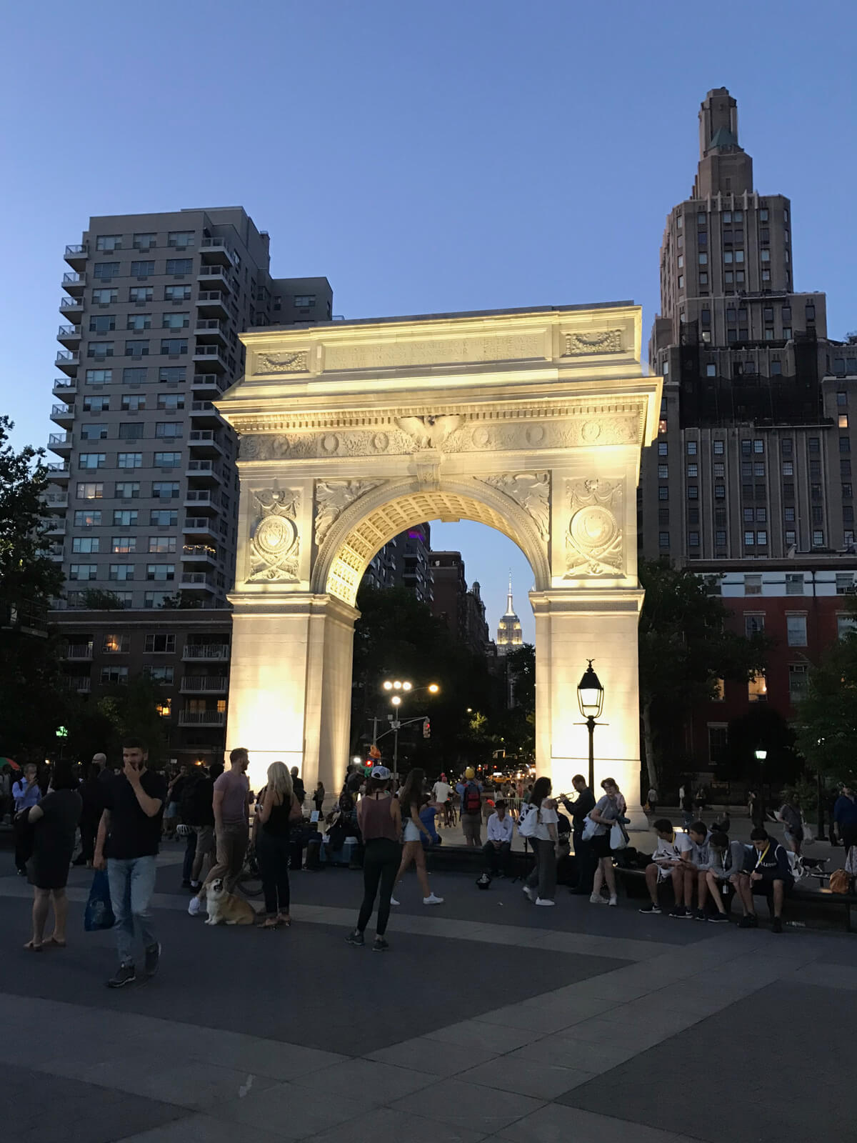 A stone arch structure lit up at night, with many people surrounding it and some taller buildings in the background