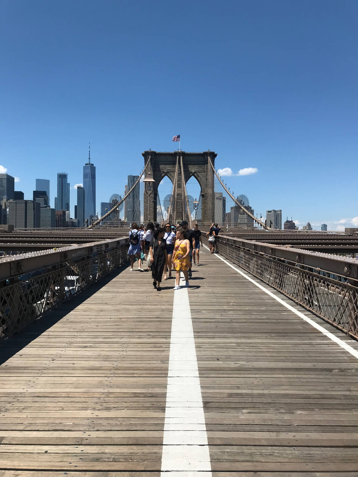 A view walking across a bridge, with an American flag on top of a pylon. Some pedestrians are walking down the bridge and conversing with each other