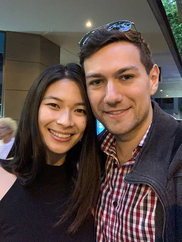 A man and woman smiling. The man is wearing a checkered red and black shirt and the woman is wearing a black halter dress