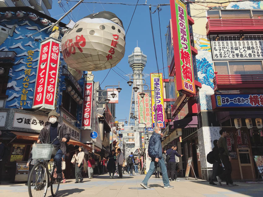 A walkway in Shinsekai with a lot of signage, taken from a low angle with the Tsutenkaku Tower in the background