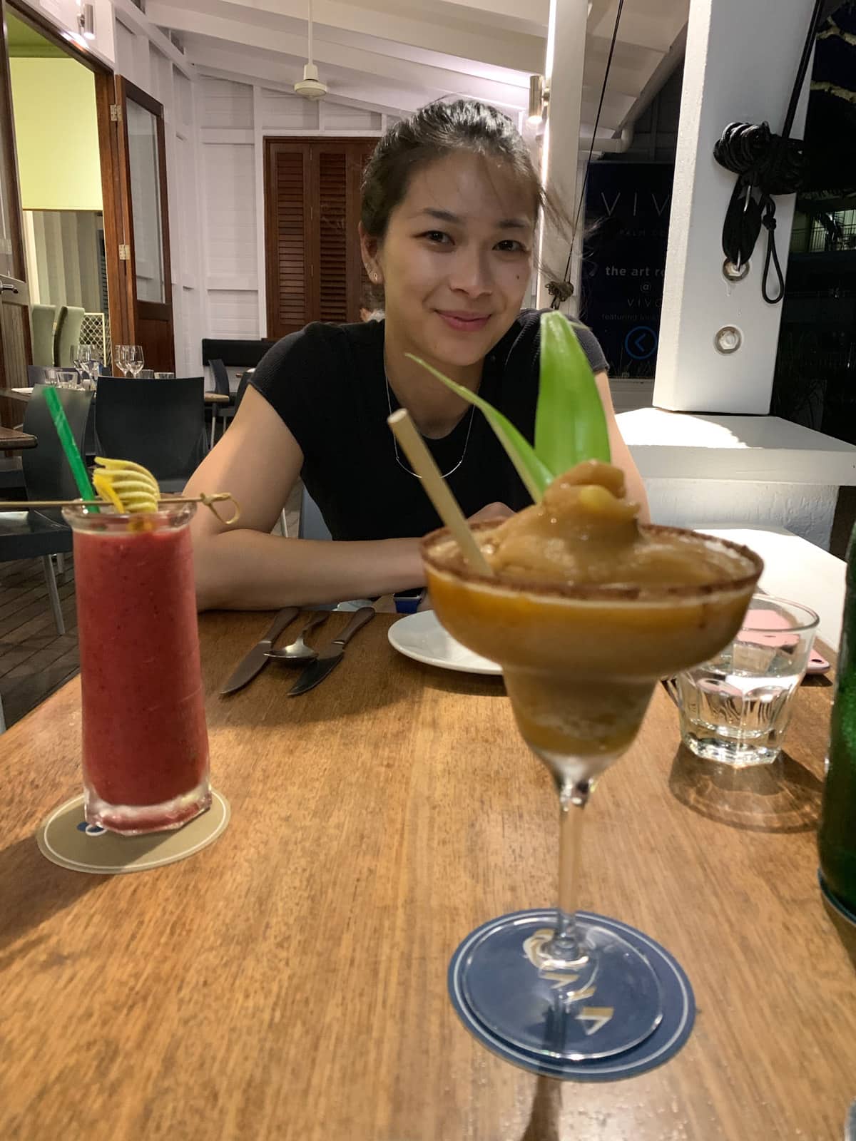 A woman sitting across from a table with her arms slightly folded. She is smiling. On the table are two cocktails and some cutlery and plates