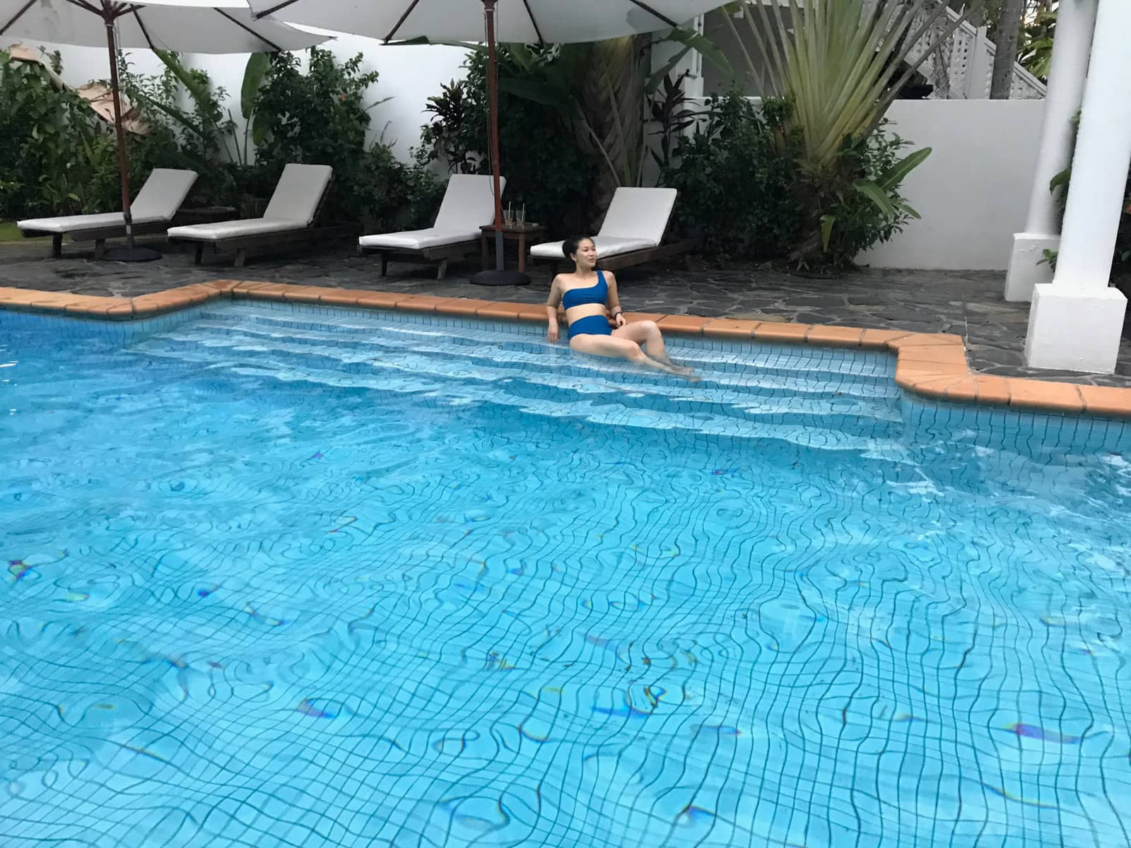 A swimming pool, on the far end are sun beds and partially in the water is a woman in a blue bikini, lying partially on her side. She is looking away from the camera
