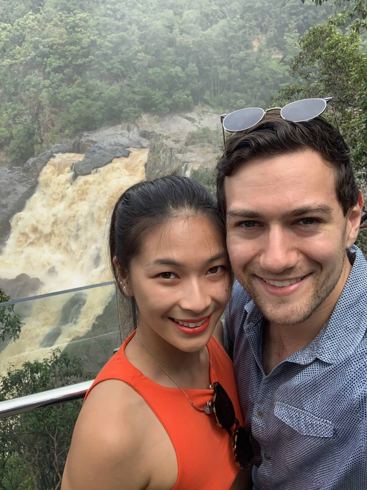 A selfie of a man and woman smiling. The woman is wearing an orange top and the man is wearing a pattered blue collared shirt and has sunglasses on top of his head. There is a waterfall in the background.