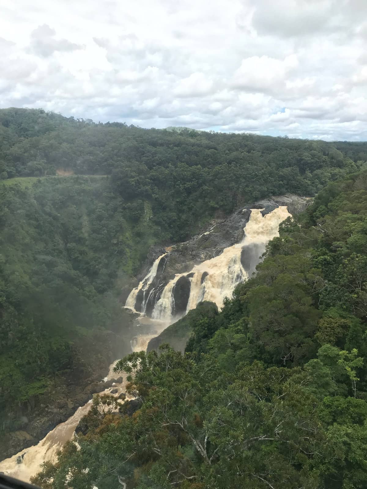 A very dirty waterfall with light brown water, seen from a high vantage point with lots of trees. There are lots of large stone faces in the waterfall’s flow