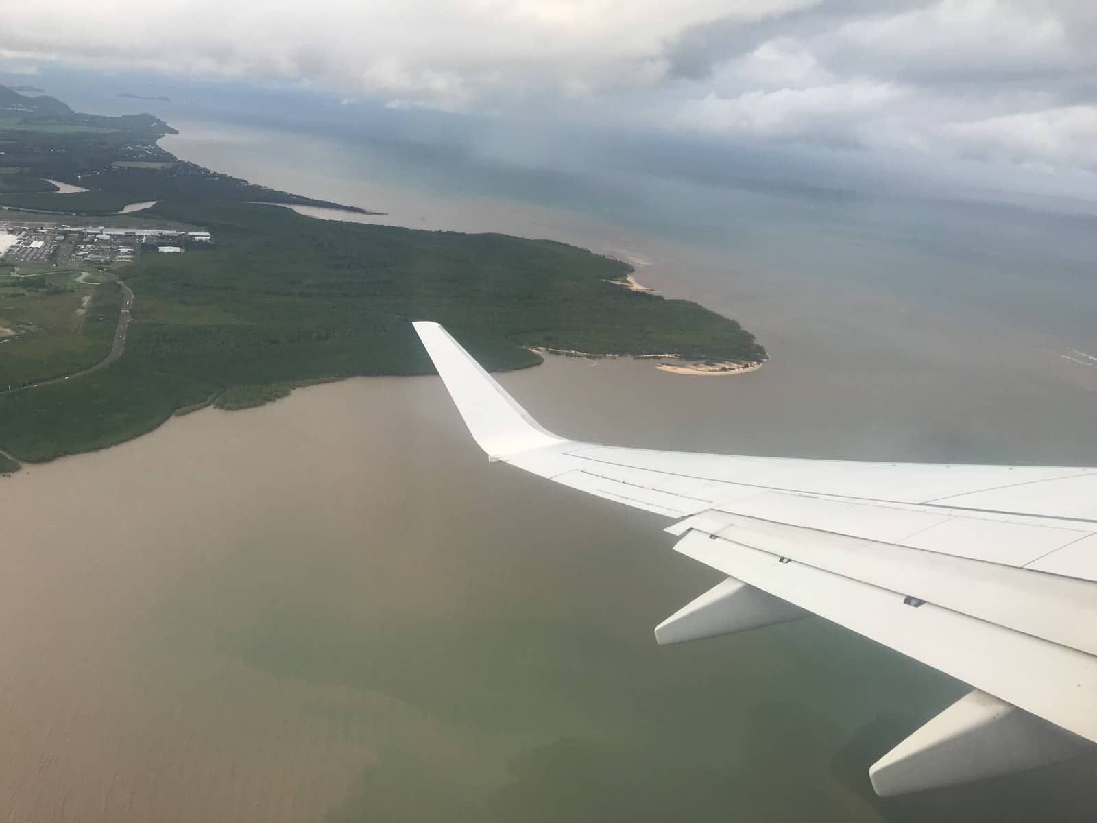 A view from a plane window of the left plane wing. It’s a very cloudy day and a green part of the coast can be seen below, as well as the ocean, which looks like a cloudy brown