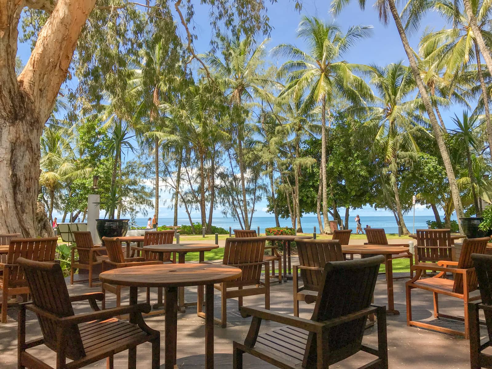An outdoor dining area with wooden chairs and tables. In the background is a beach, the ocean, and many palm trees