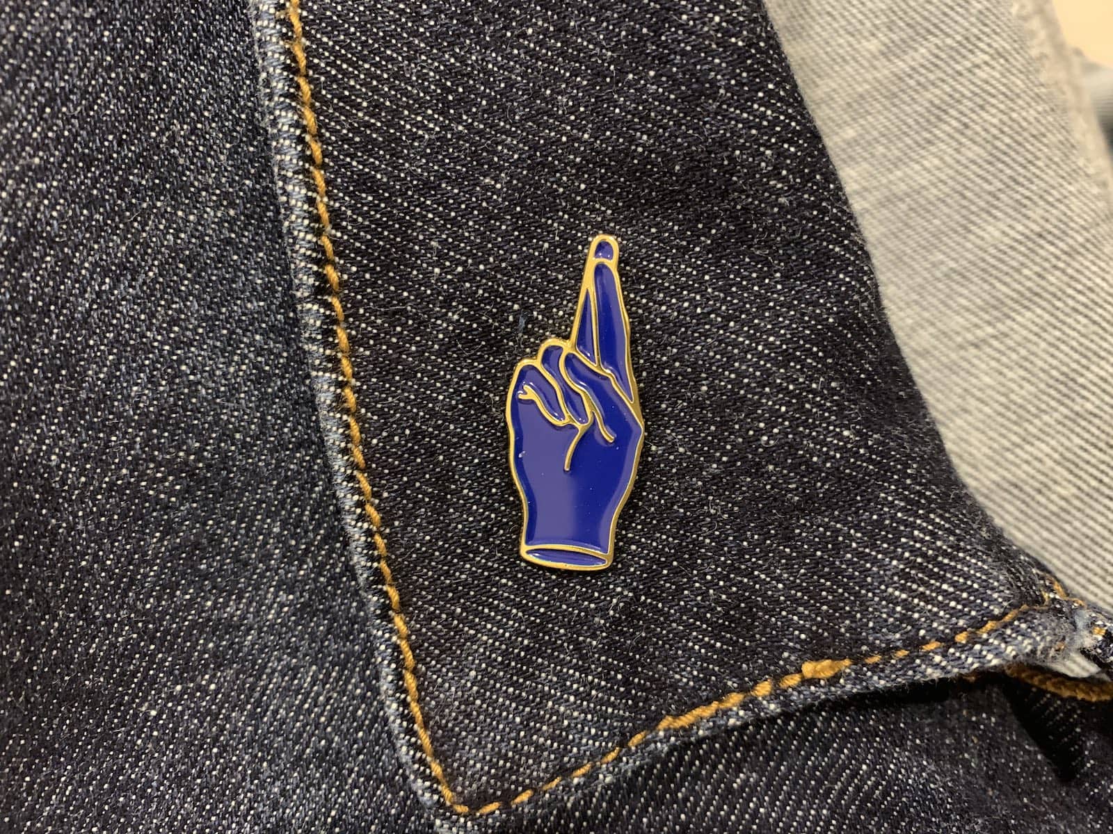 A blue enamel pin of the fingers crossed sign, on a dark denim collar