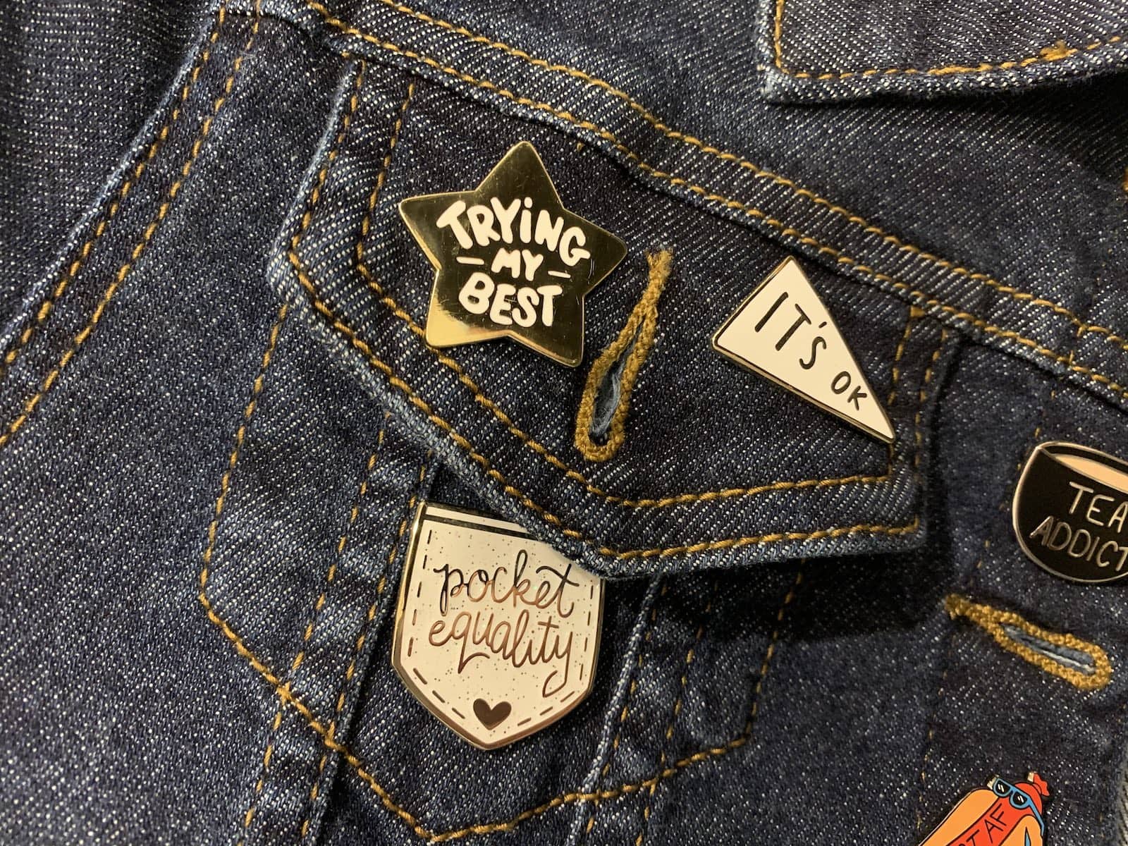 Part of a dark denim jacket with two pins in view: a pennant flag reading “It’s OK” and a star reading “Trying my best”.