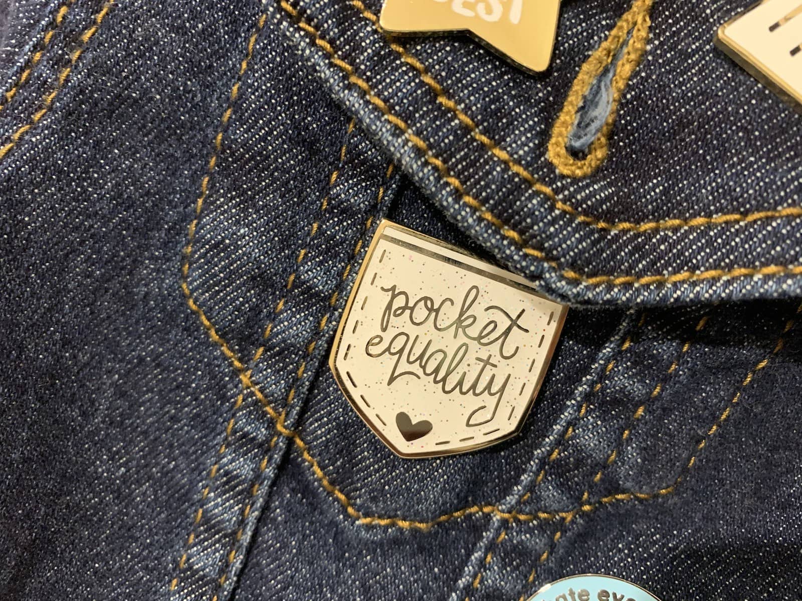 A white enamel pin shaped like a pocket, reading “pocket equality”, pinned on the pocket of a denim jacket