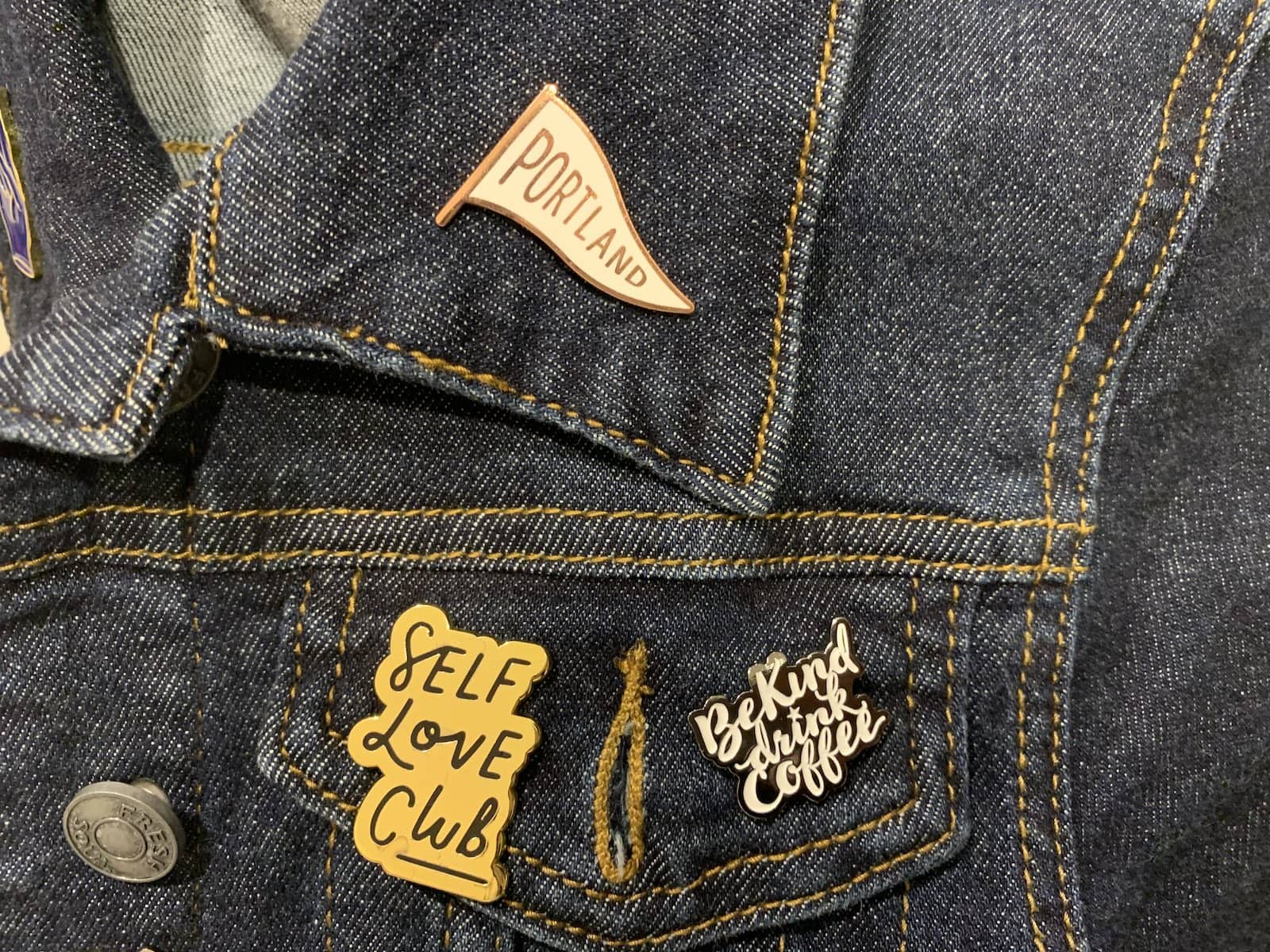 A dark denim jacket with pins reading “Portland”, “Self-love club” and “Be kind, drink coffee”.