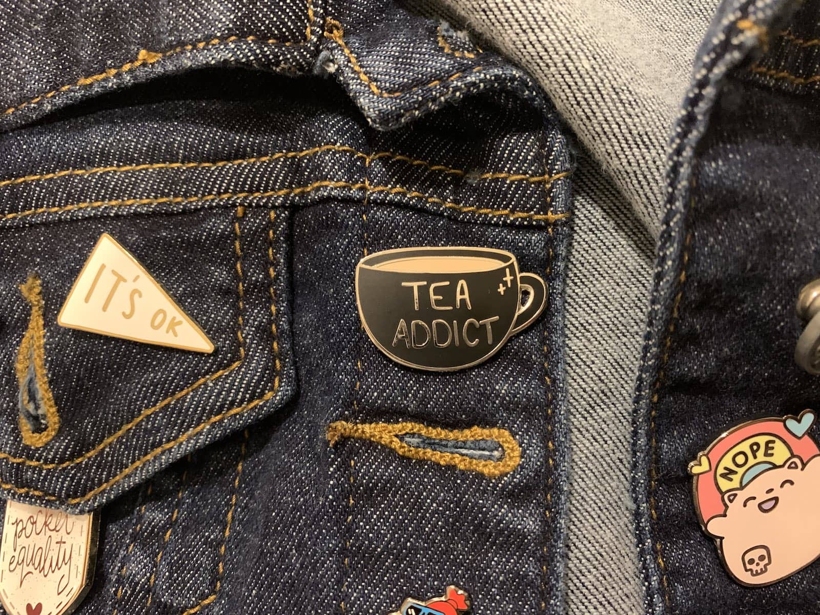 A black enamel pin of a round bottom teacup with the writing “Tea Addict” on it; it’s pinned on dark denim