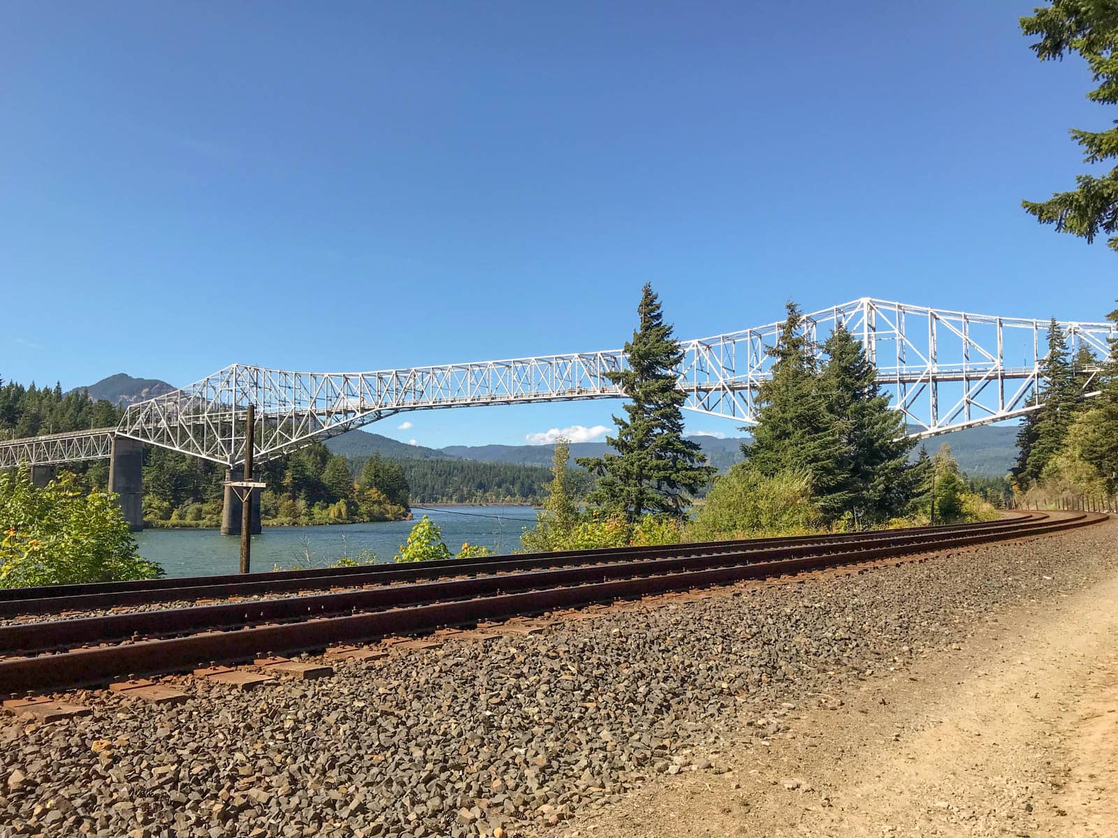 A white structured bridge across a gorge, seen from the side of railway tracks on one side of the bridge. It is a bright day with very blue skies.