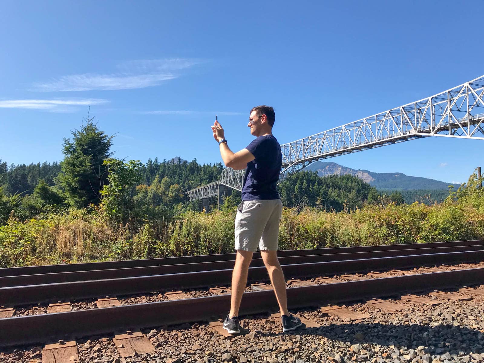 A man wearing sunglasses, taking a photo on a smartphone. There is a gorge and bridge going across it on his left. He is standing in front of some train tracks.