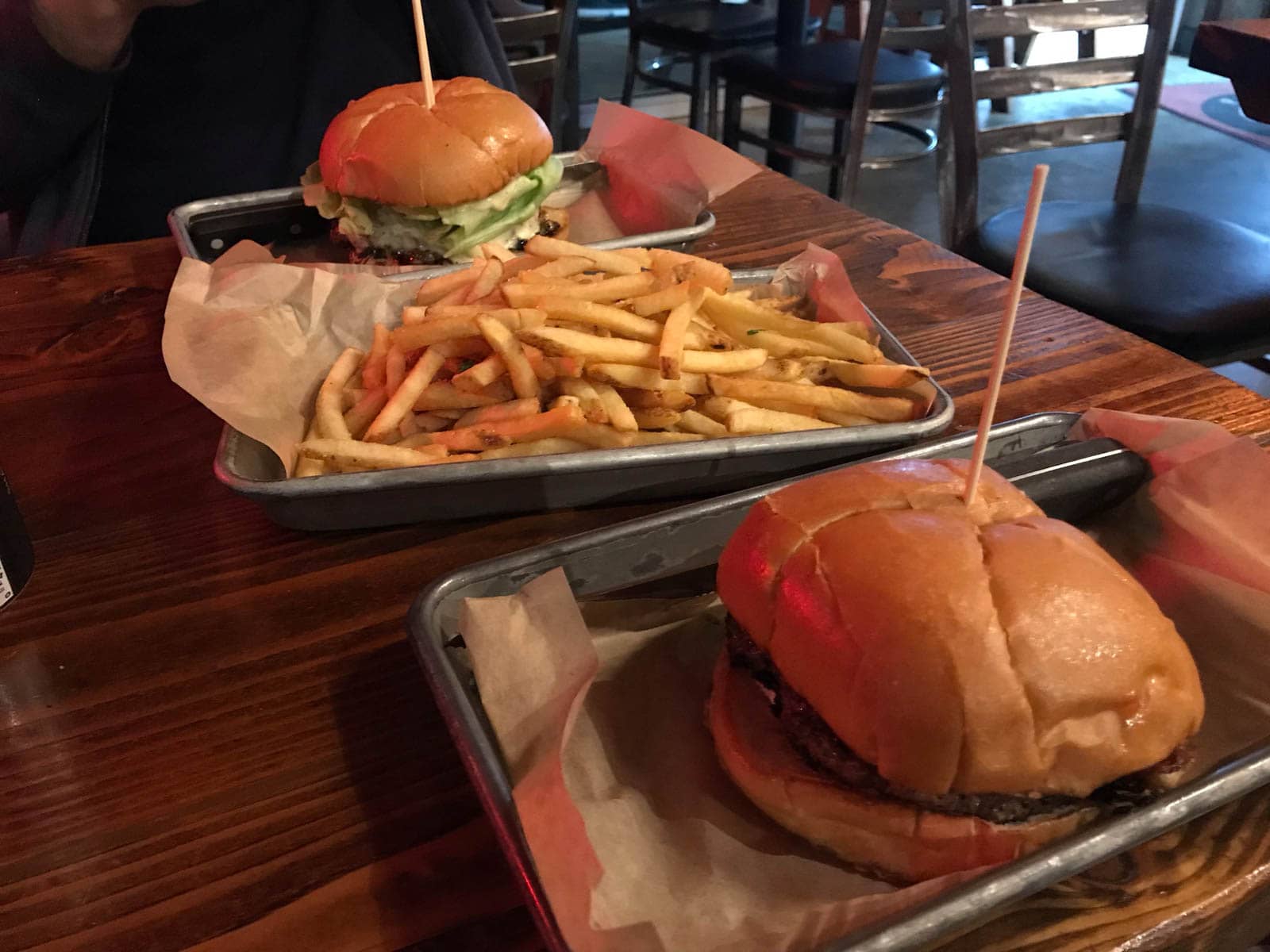 Three silver trays on a wooden table. Two of the trays have burgers in them, held together with a skewer. The other tray in the middle of the table has a load of fries on it.