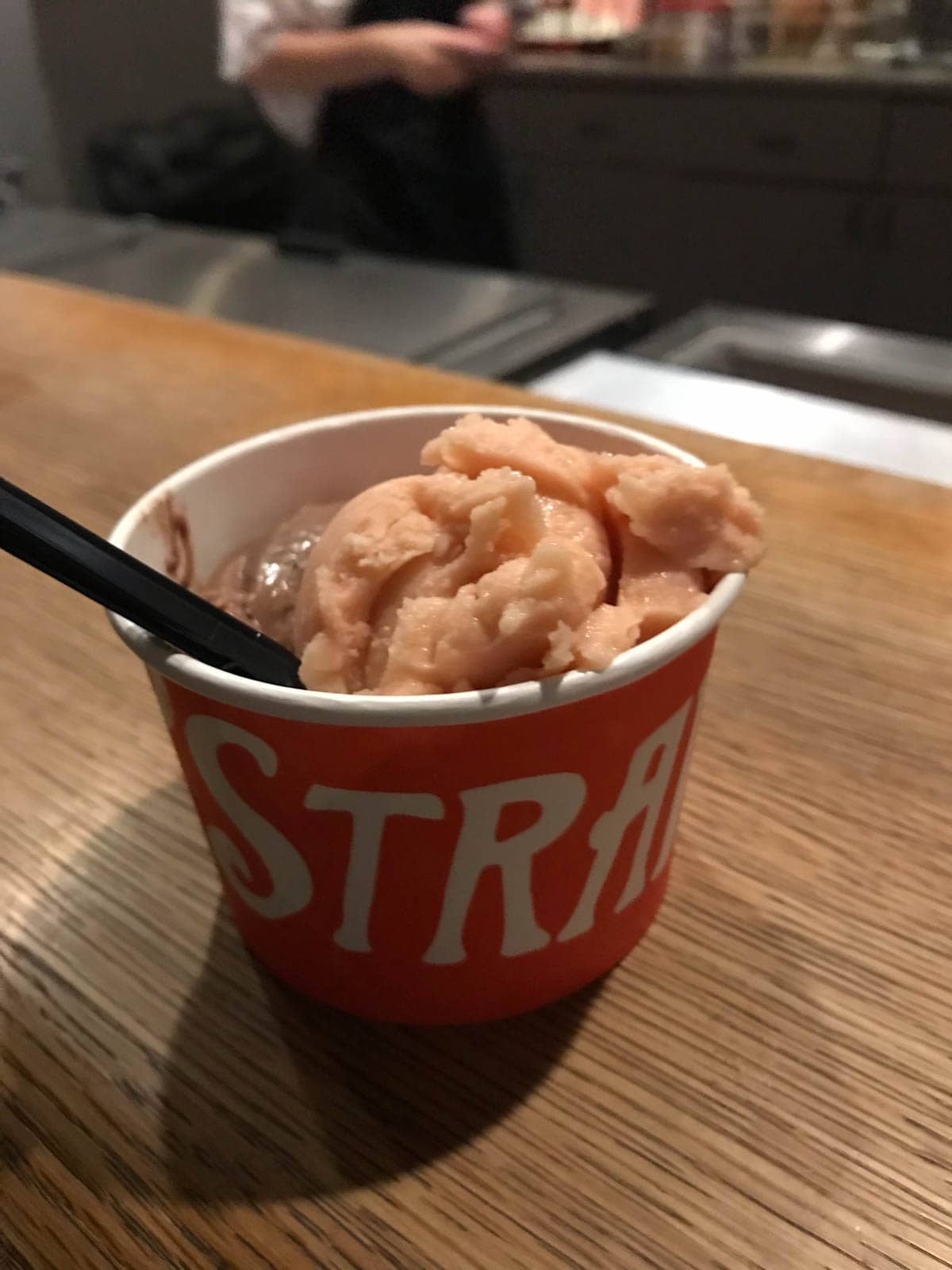 A paper cup with pink ice cream in it. A black spoon sticks out of the cup.