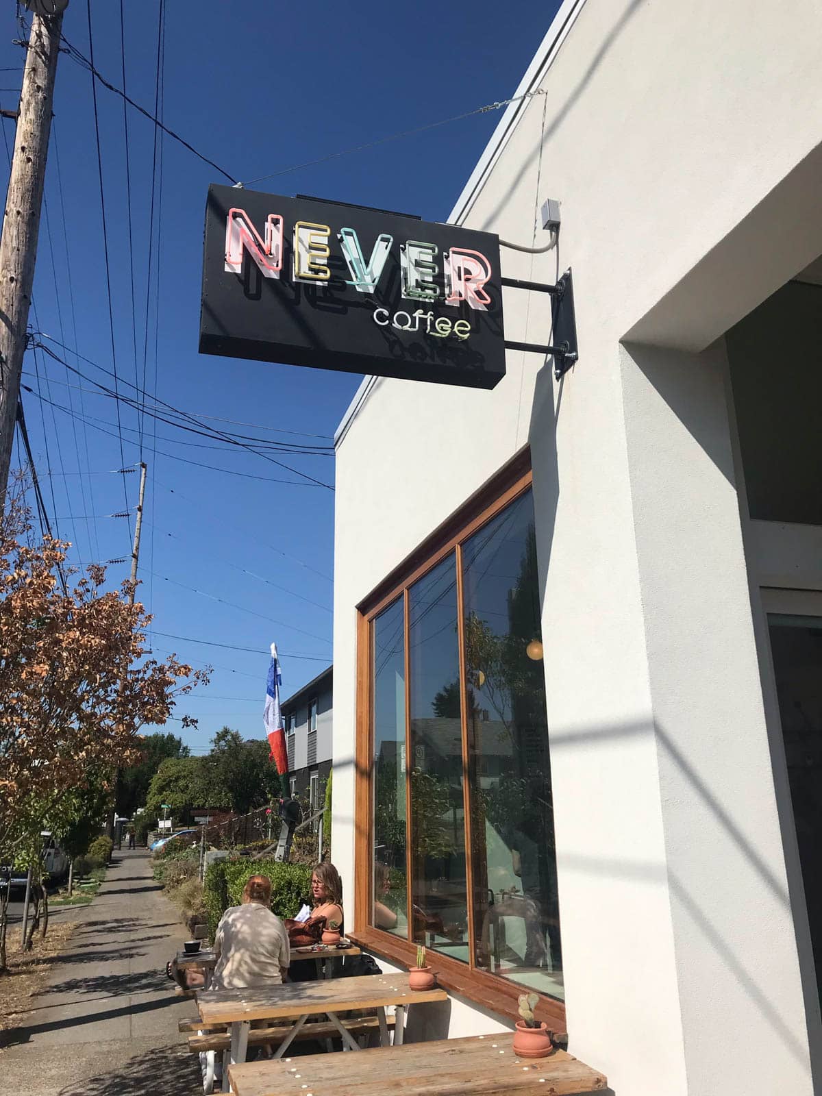 The front of a coffee shop, with a neon sign reading “Never coffee”. Two people sit outside the coffee shop at a wooden table.