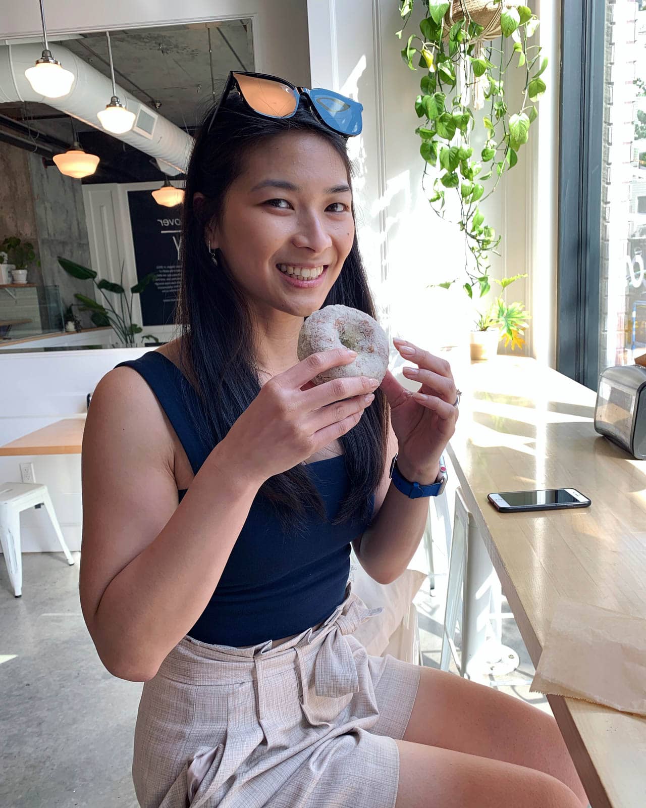 A woman with dark hair and sunglasses on top of her head, grinning and holding a light coloured doughnut. She is sitting at a high table by a window.